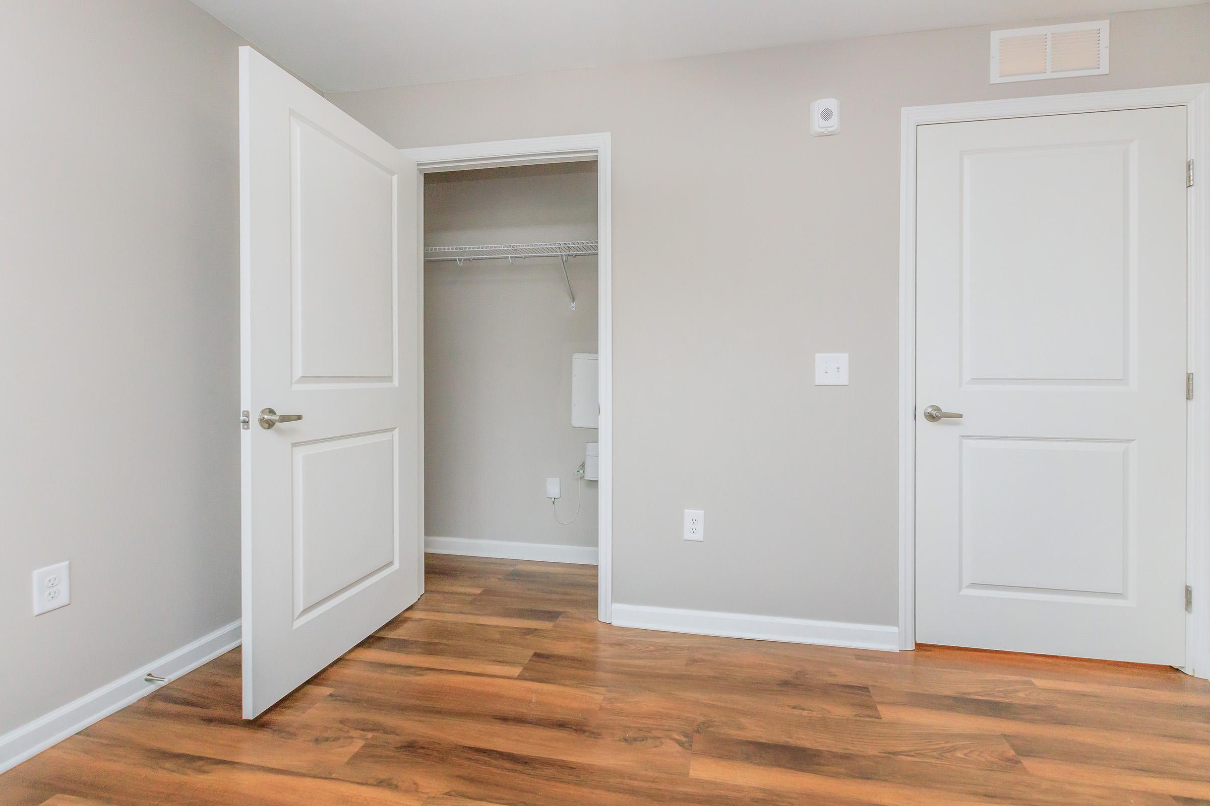 A spacious, well-lit room featuring two open doors. One door leads to a small closet, while the other may lead to a bathroom or another space. The walls are painted a neutral light gray, and the flooring is wood laminate. A thermostat is visible on one wall.