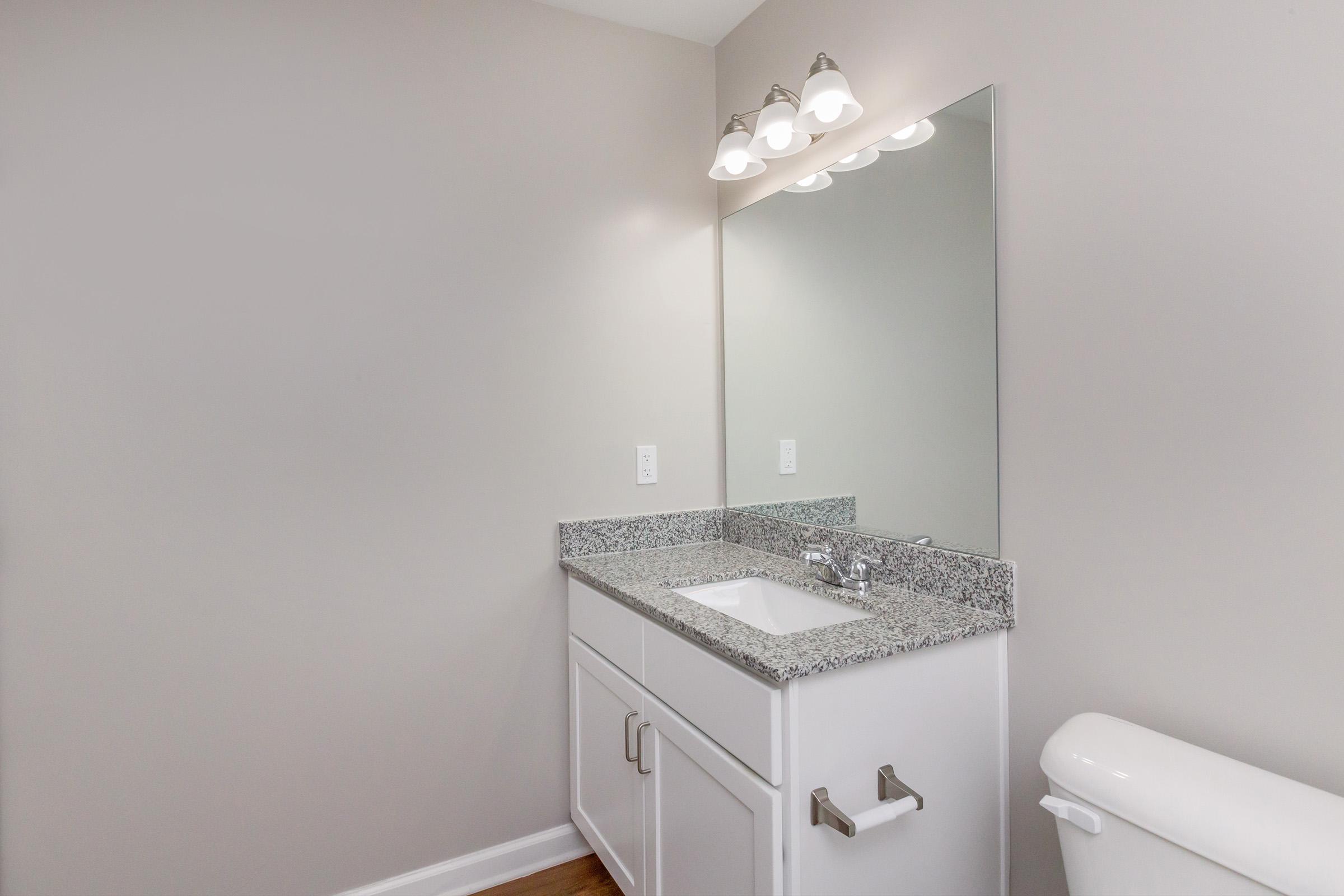 A modern bathroom featuring a light gray wall, a granite countertop with a sink, a faucet, and a large mirror above. Three light fixtures are mounted above the mirror, and a white toilet is visible to the right. The overall design is clean and minimalist.