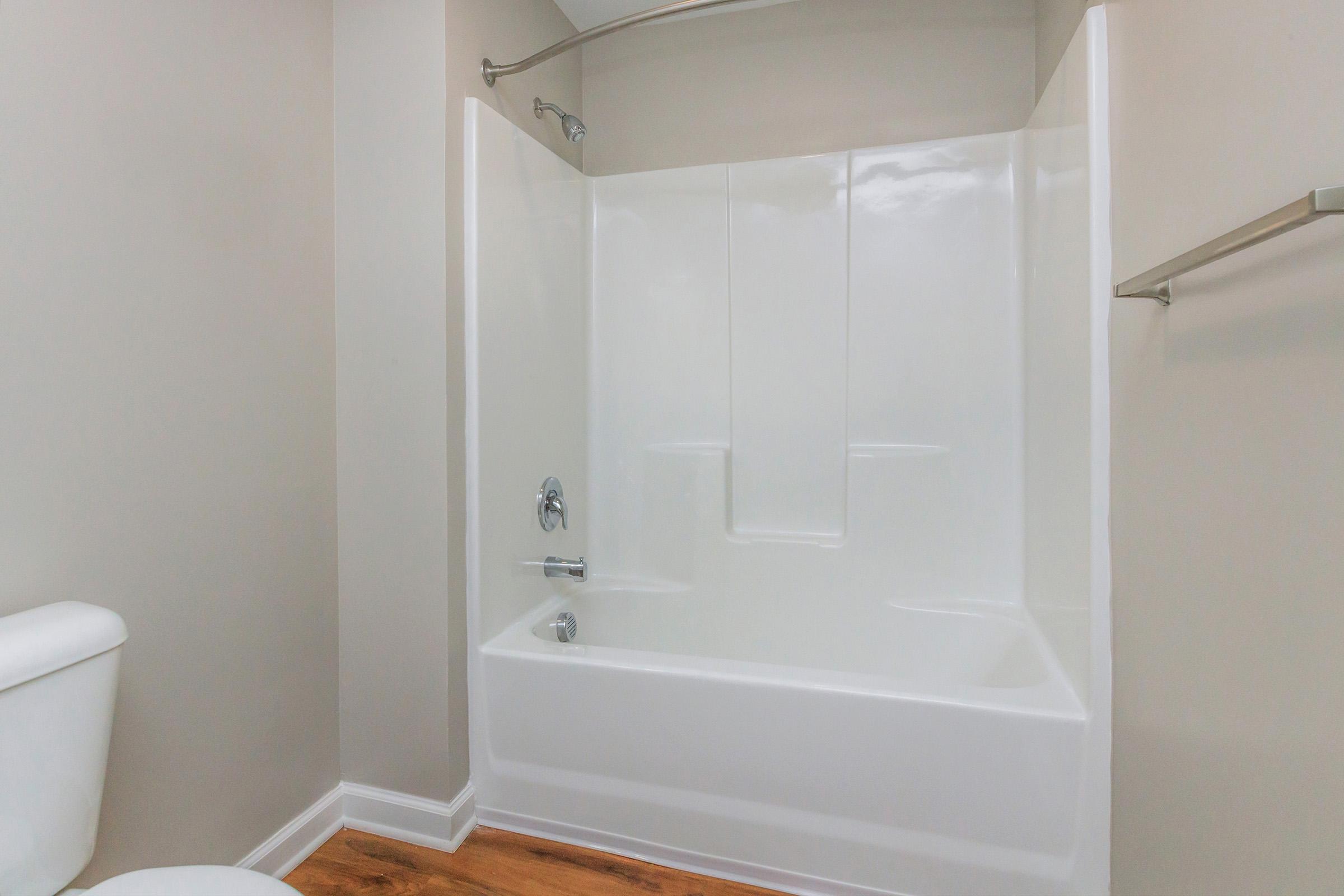 A clean and modern bathroom featuring a white bathtub with a showerhead, beige wall paint, and wooden flooring. There is a white toilet to the left and a towel bar on the right, contributing to a simple and streamlined design.