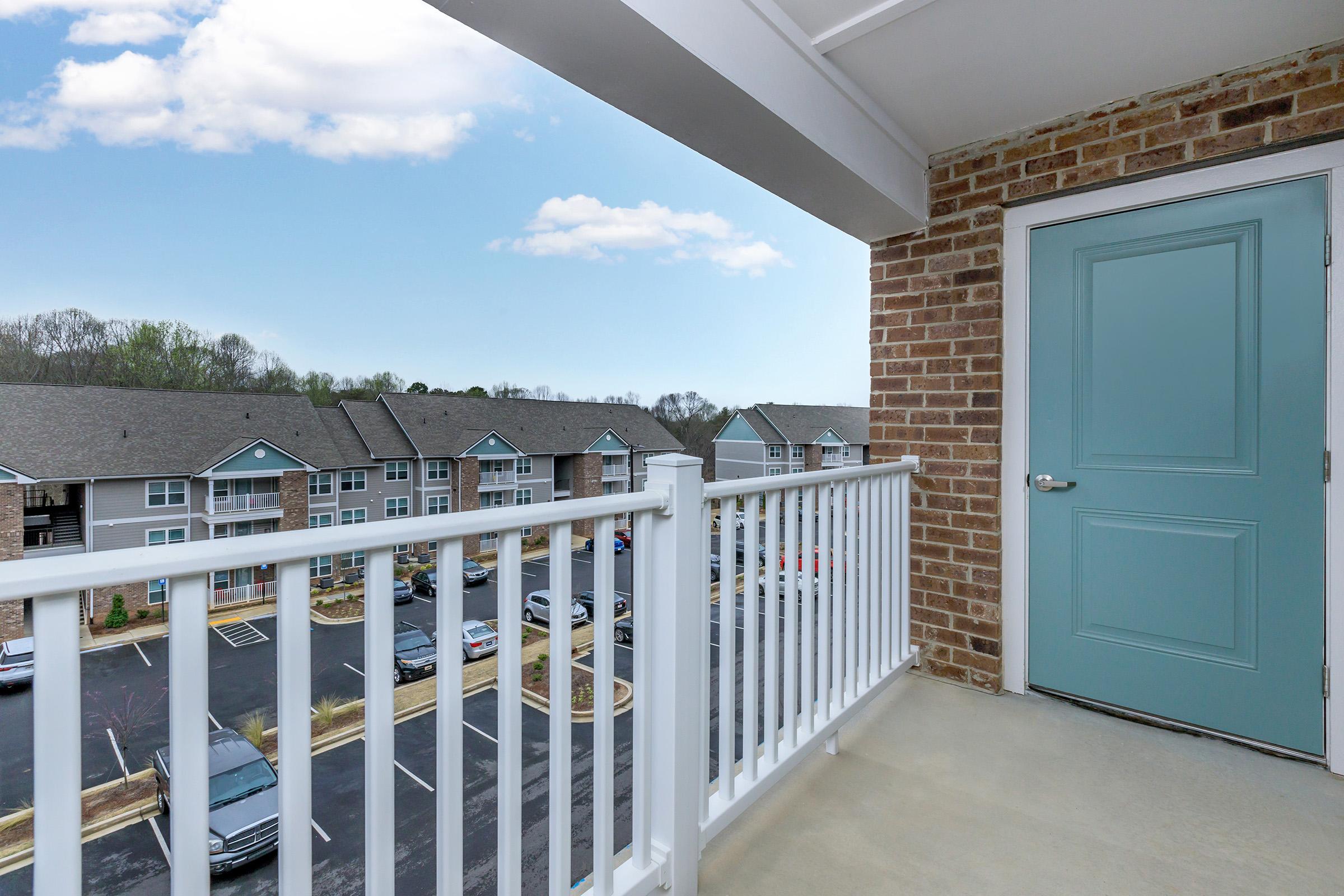 View from a balcony overlooking a parking lot and residential buildings. The clear sky features a few clouds, and a turquoise door is visible in the background.