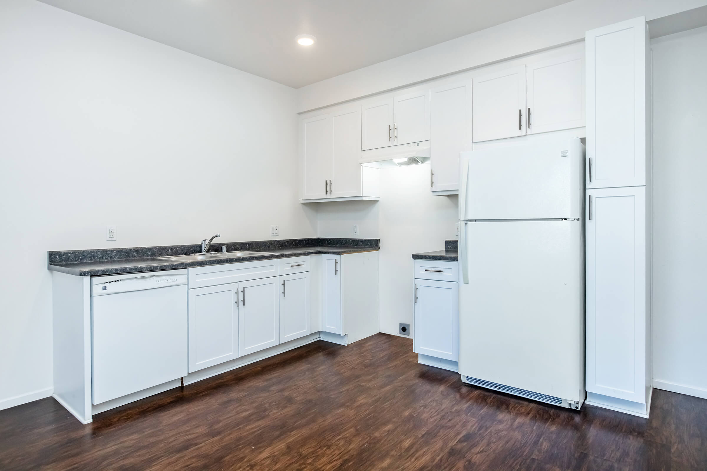 A modern kitchen featuring white cabinetry, a white refrigerator, and a dark countertop. The kitchen has a spacious layout with a sink and a dishwasher integrated into the cabinetry. The floor is made of dark wood, and the walls are painted in a light color, contributing to a bright and clean aesthetic.
