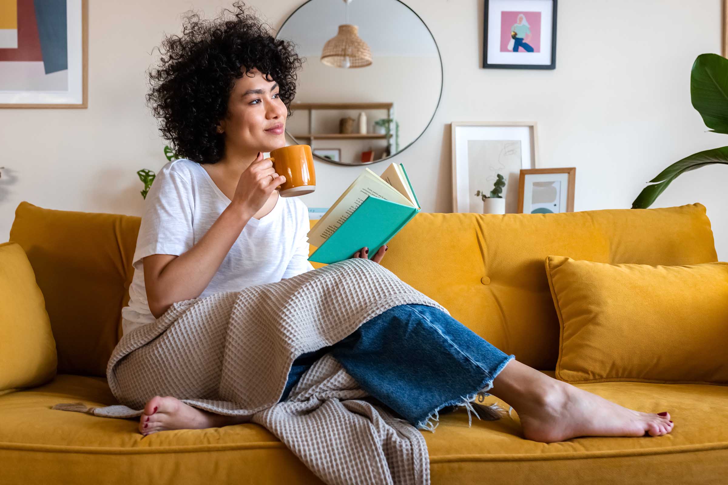 A young woman with curly hair sits comfortably on a mustard yellow sofa, wrapped in a blanket. She holds a mug in one hand and reads a book with the other. Decorative elements, like framed artwork and plants, add a cozy ambiance to the living space.