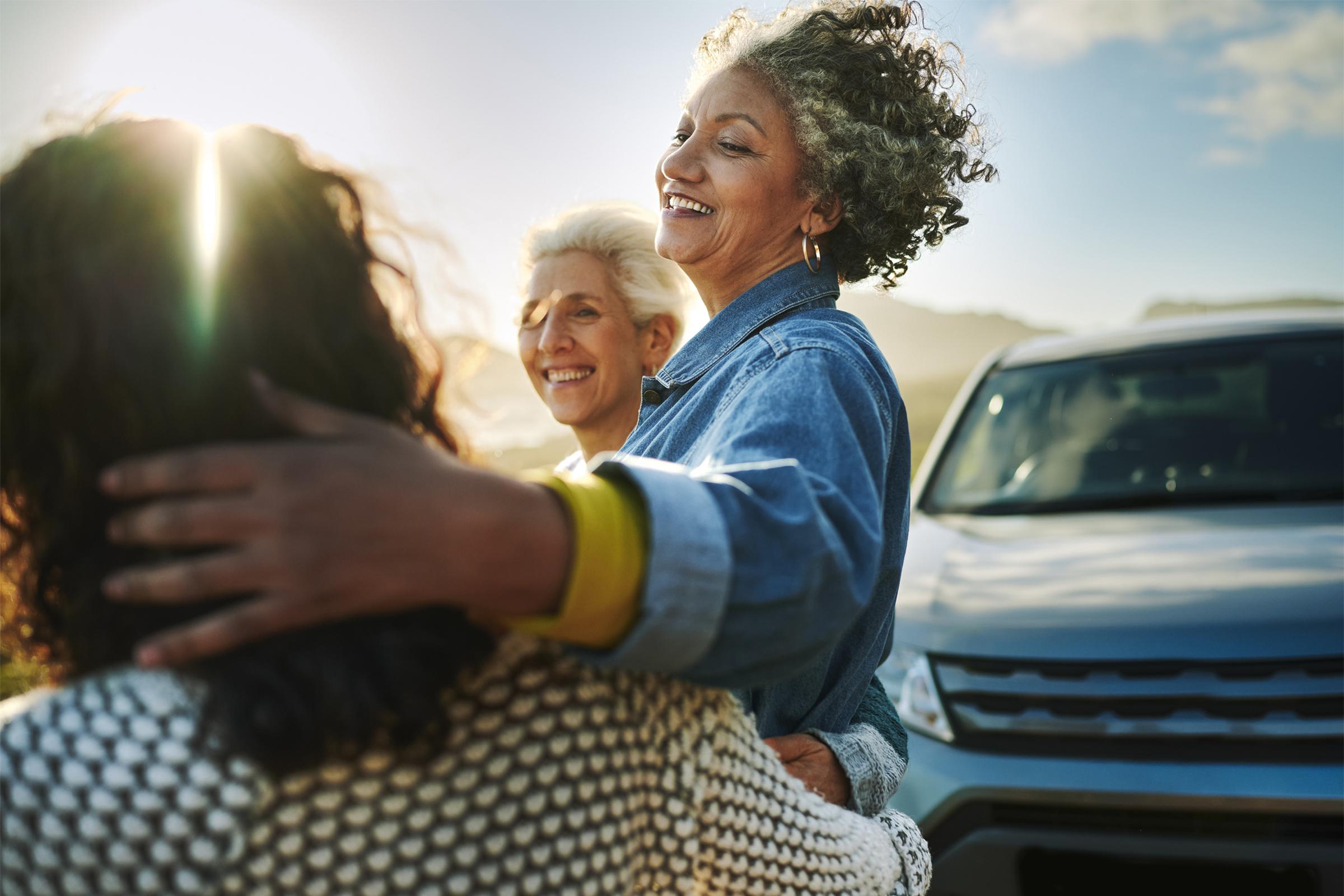 Three women enjoying a joyful moment outdoors, hugging and smiling. The sun shines in the background, creating a warm glow. One woman with curly hair is lifted by another, while a third woman watches with a smile, with a car parked nearby. The setting suggests a friendly gathering or reunion.
