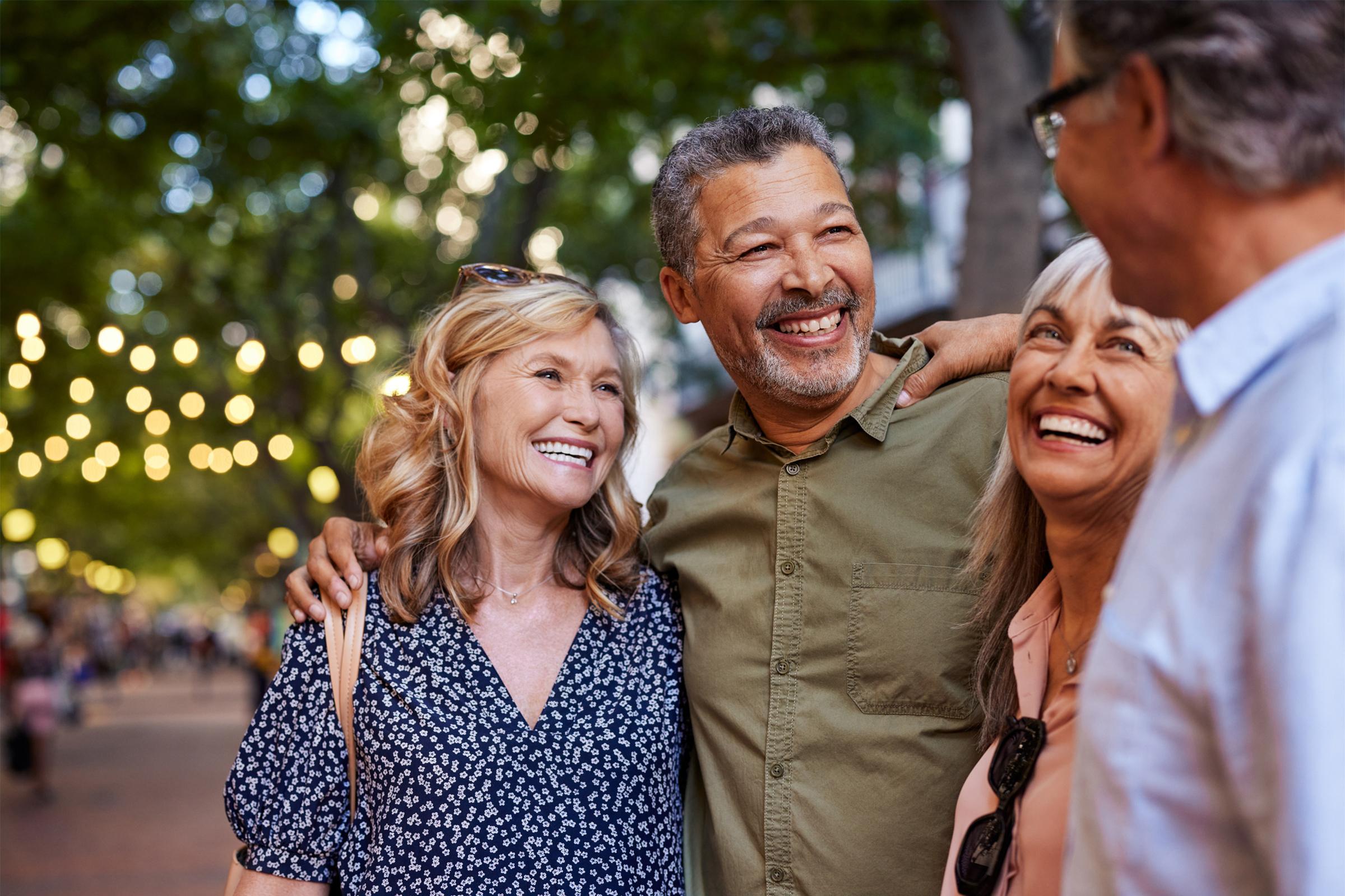 A group of four smiling adults enjoying a casual moment outdoors, surrounded by greenery and warm ambient lighting from string lights. They appear to be friends, laughing and sharing a happy conversation, conveying feelings of joy and companionship.