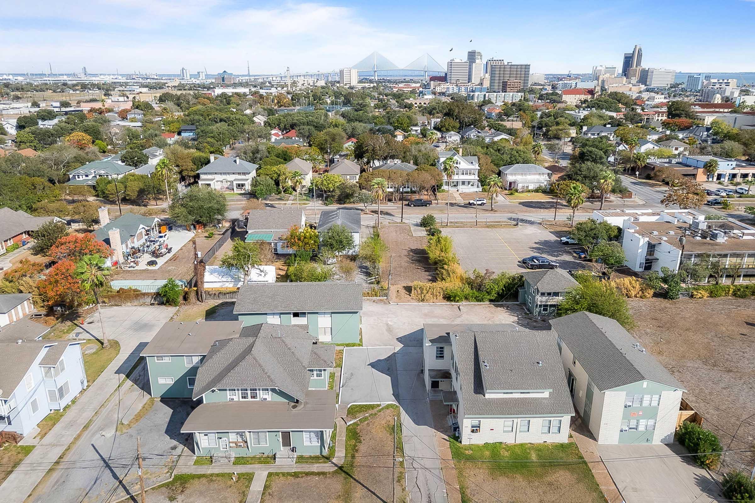 Aerial view of a residential neighborhood featuring a mix of single-family homes and apartment buildings. The image shows greenery, roads, and nearby urban structures, with a skyline visible in the background. The scene conveys a blend of suburban and urban environments.