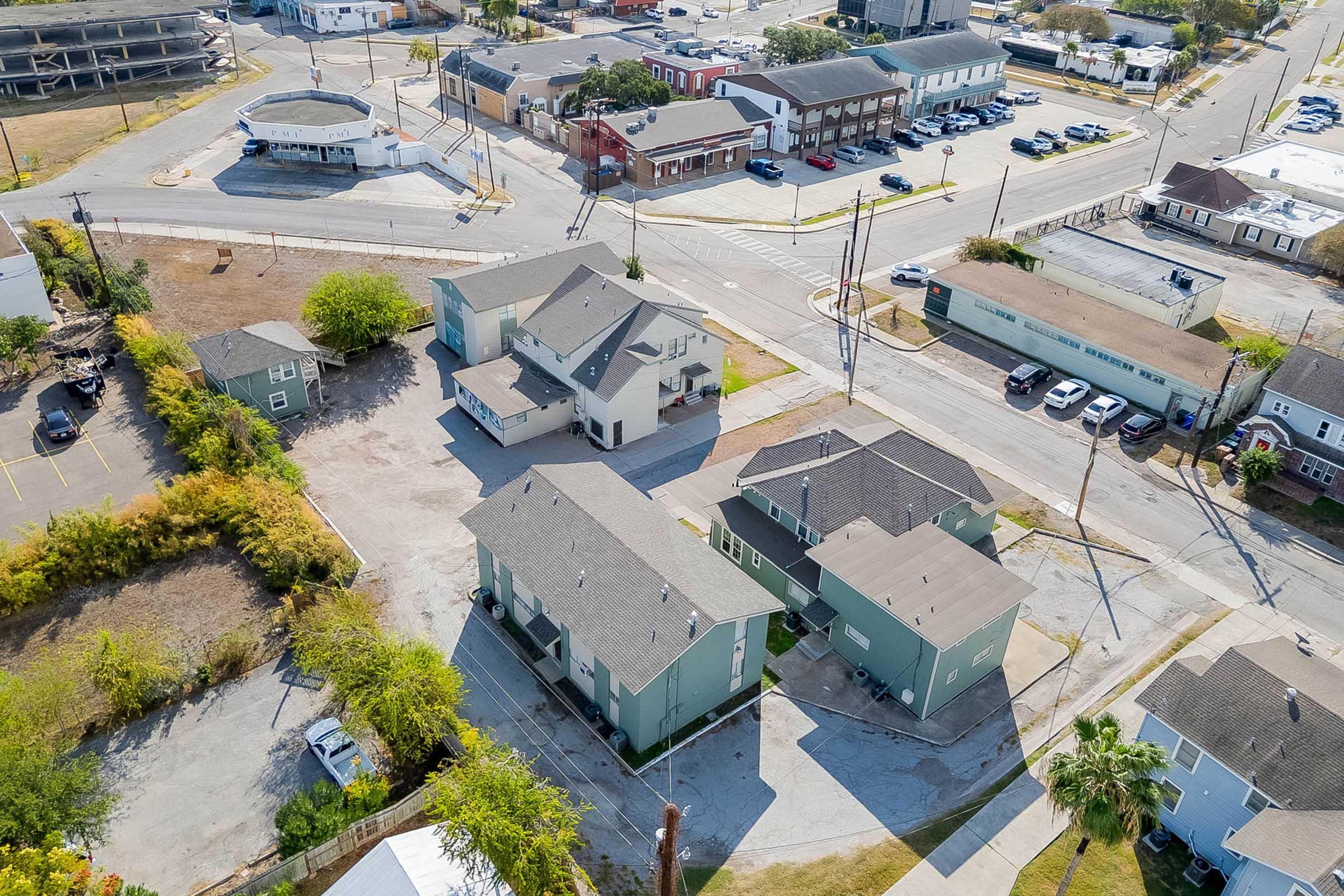 Aerial view of a neighborhood featuring various residential buildings, including multi-story houses and smaller structures. Surrounding streets and parked vehicles are visible, along with green trees and open land. The scene depicts a blend of urban and suburban elements.