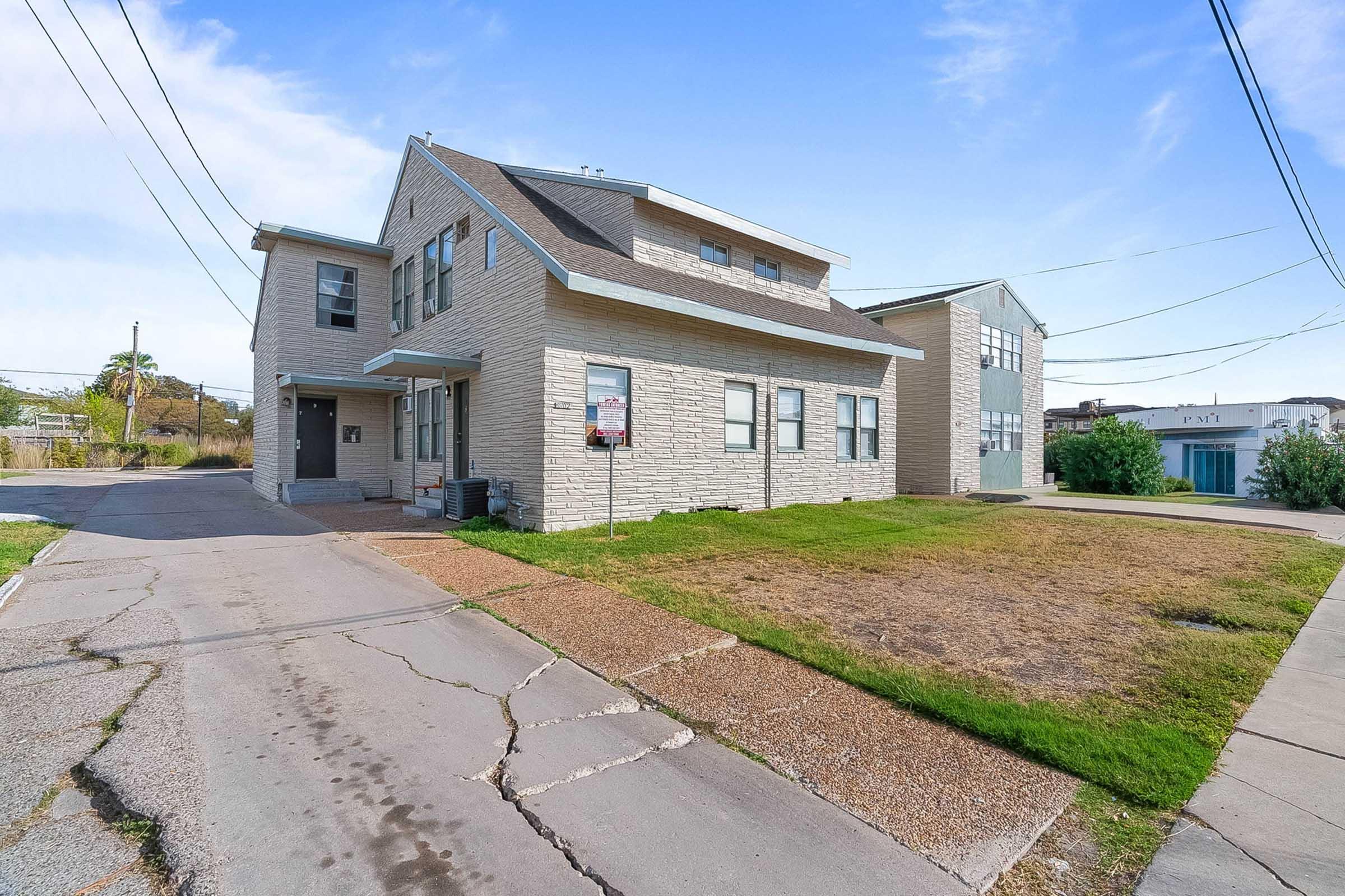 A two-story light-colored stone building situated on a grassy area with a gravel driveway. Power lines run overhead, and there are neighboring buildings in the background. The setting is relatively open with a clear blue sky.