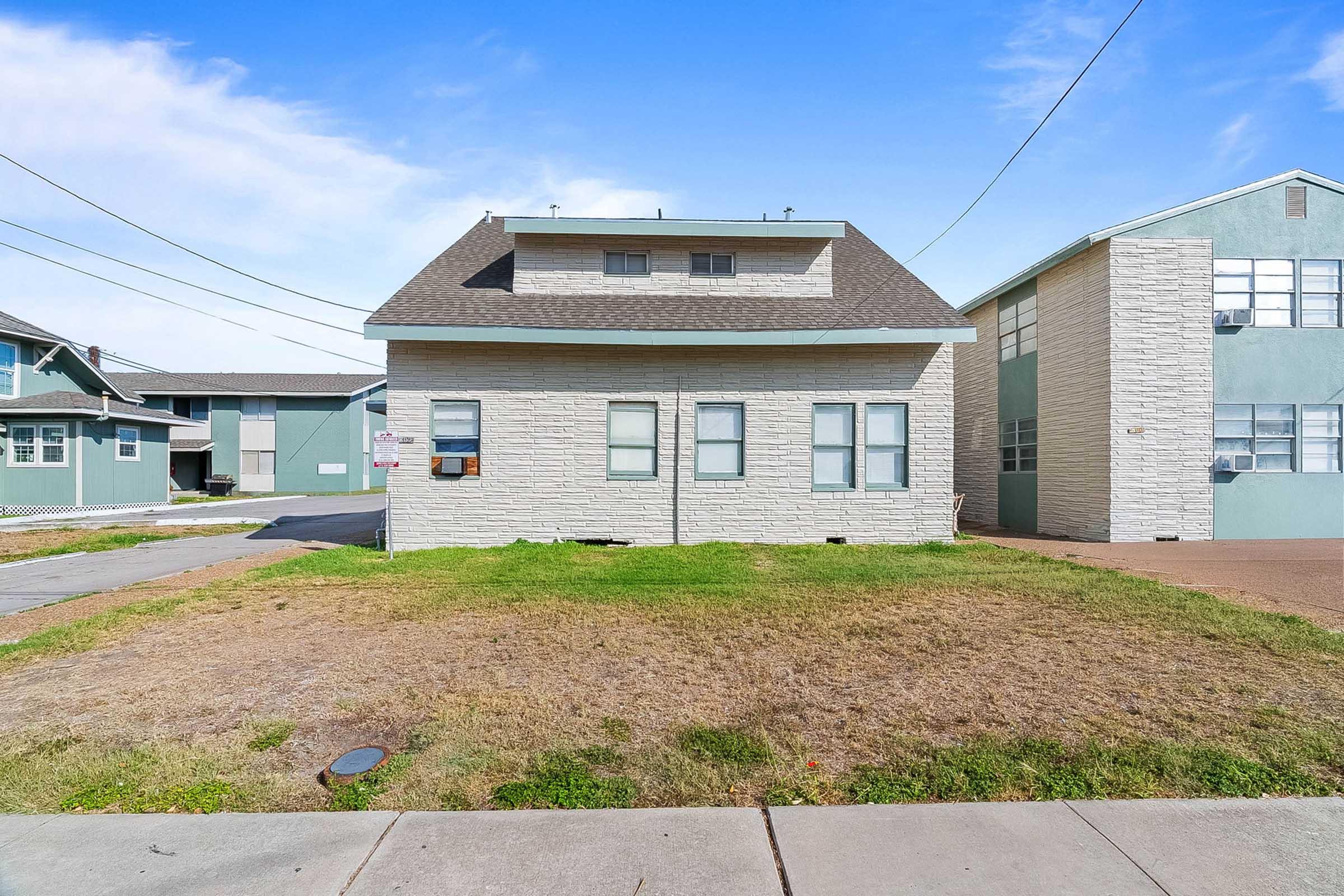 A two-story beige building with a sloped roof, featuring several windows, sits on a grassy lot. To the right, a multi-story building with a light blue façade is visible. The sky is clear with a few clouds, and power lines are overhead. The area appears residential.