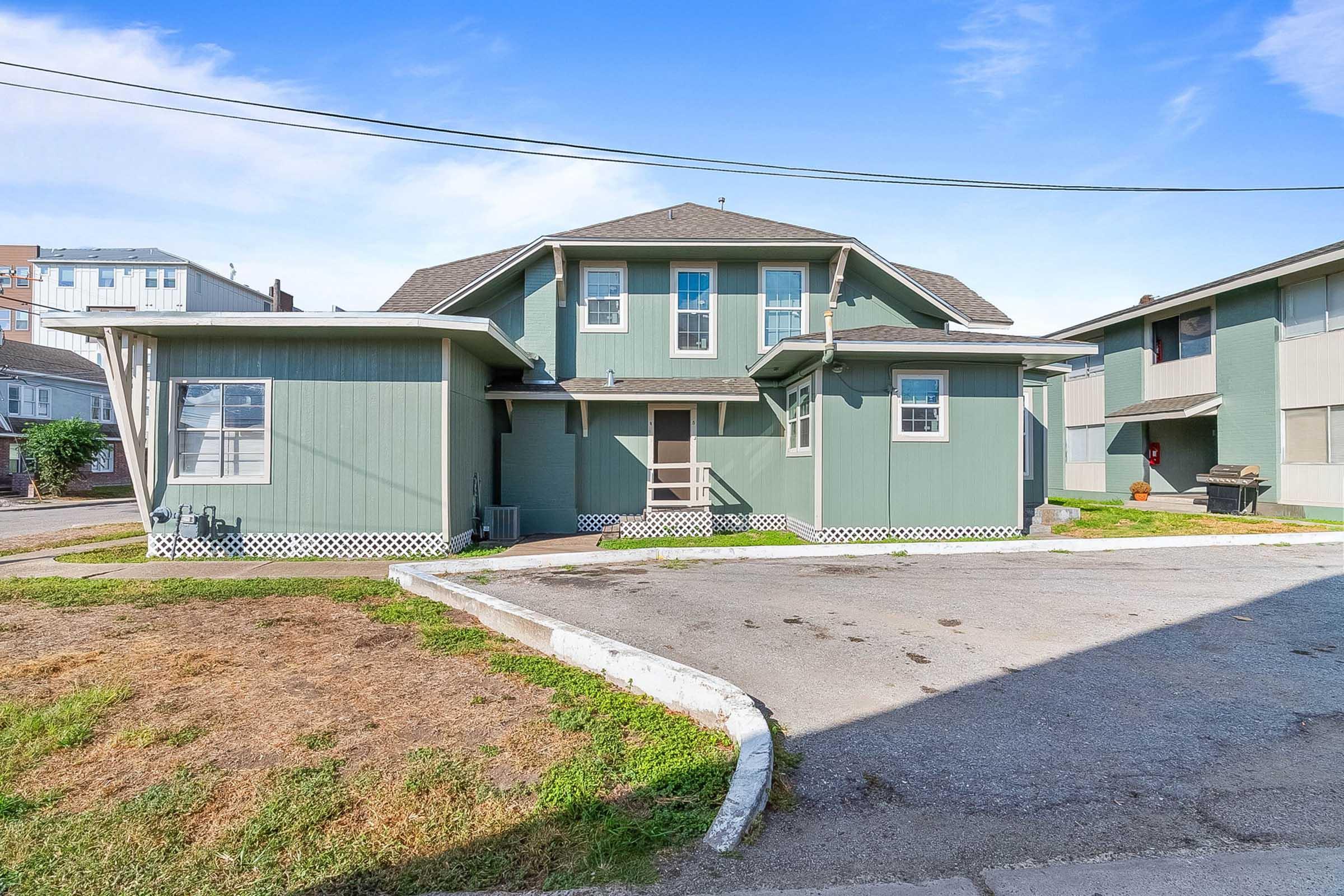 Two-story green building with a gabled roof and large windows. The structure has a porch and is surrounded by a grassy area and parking space. In the background, there are taller buildings and a clear blue sky. The scene appears well-maintained and inviting.