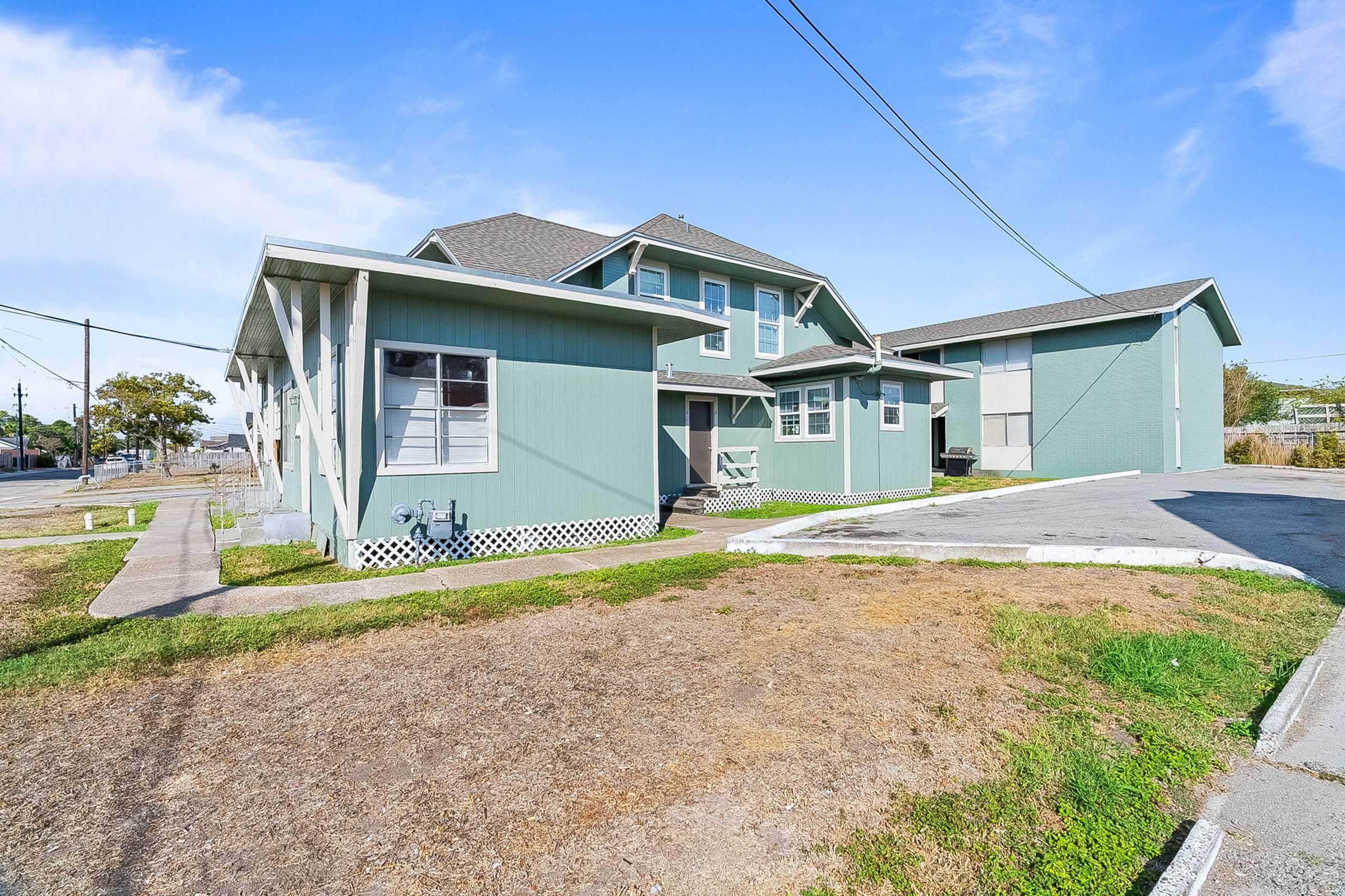A two-story green house with a gray roof and white trim. It features multiple windows and a parking area in front. The surrounding landscape includes patches of grass and pavement. Power lines are visible in the background against a clear blue sky.