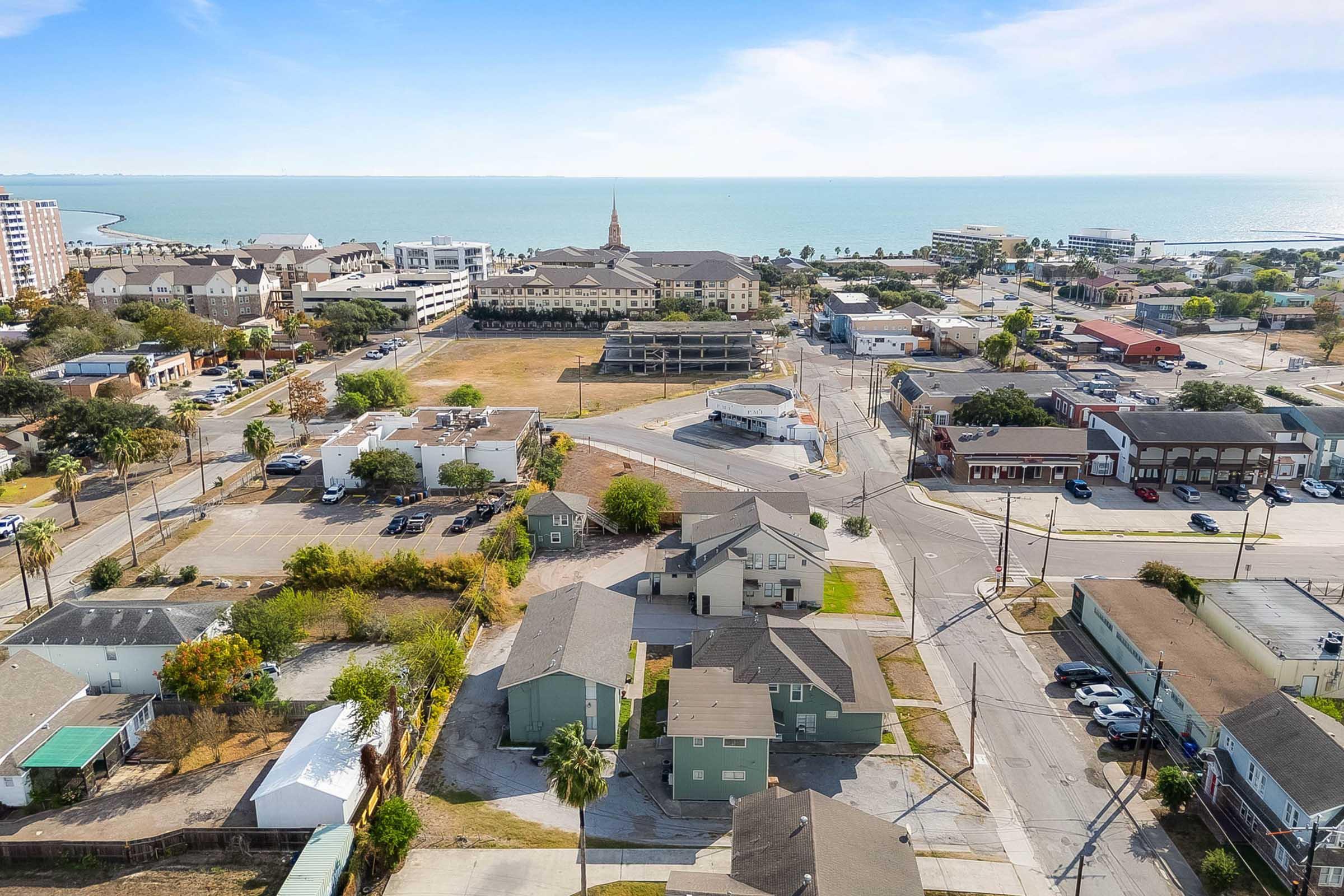  aerial view of a coastal town featuring a mix of low-rise buildings, residential homes, and businesses. In the background, the ocean can be seen, along with a distant church steeple and other structures along the waterfront. The scene captures a blend of urban and natural landscapes.