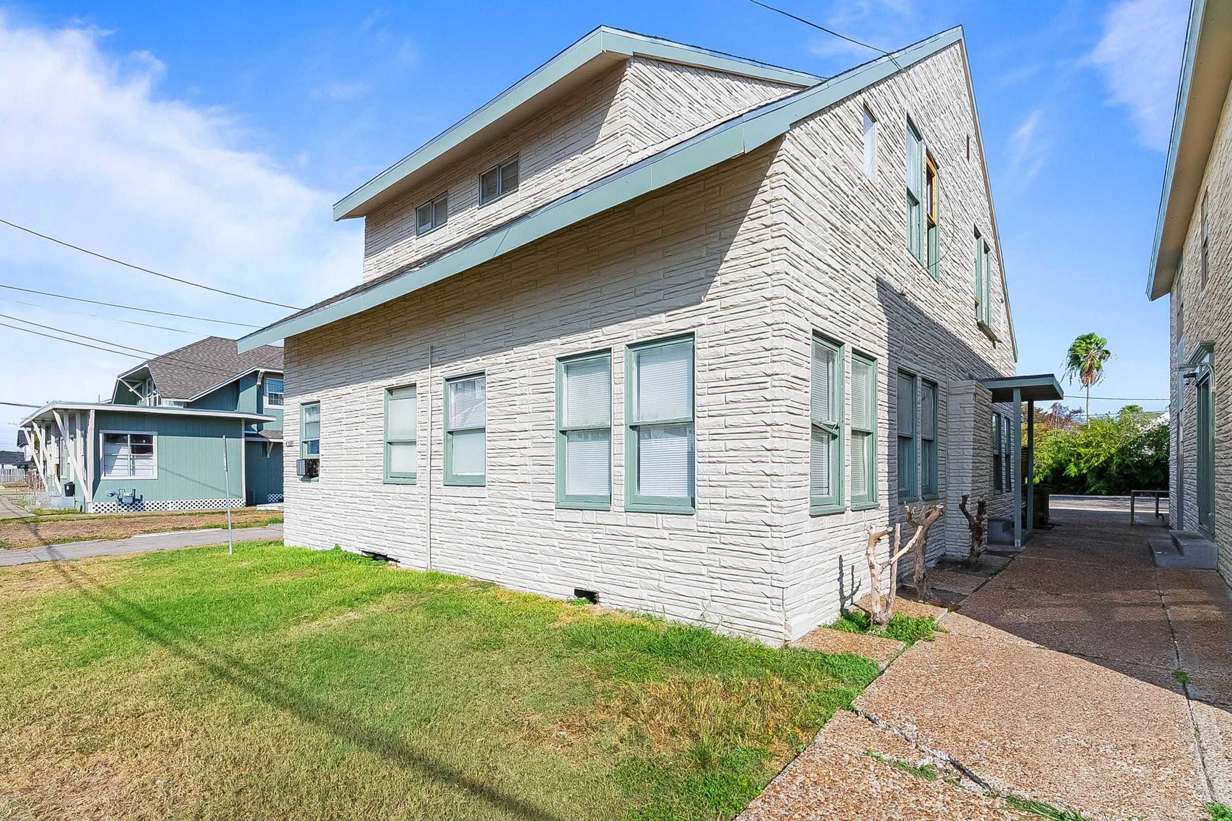 A view of the side of a two-story, beige residential building with a textured exterior. The building features multiple windows and green trim. In the foreground, there is a grassy area and a paved walkway. Power lines are visible in the background, with other buildings seen at a distance under a blue sky.