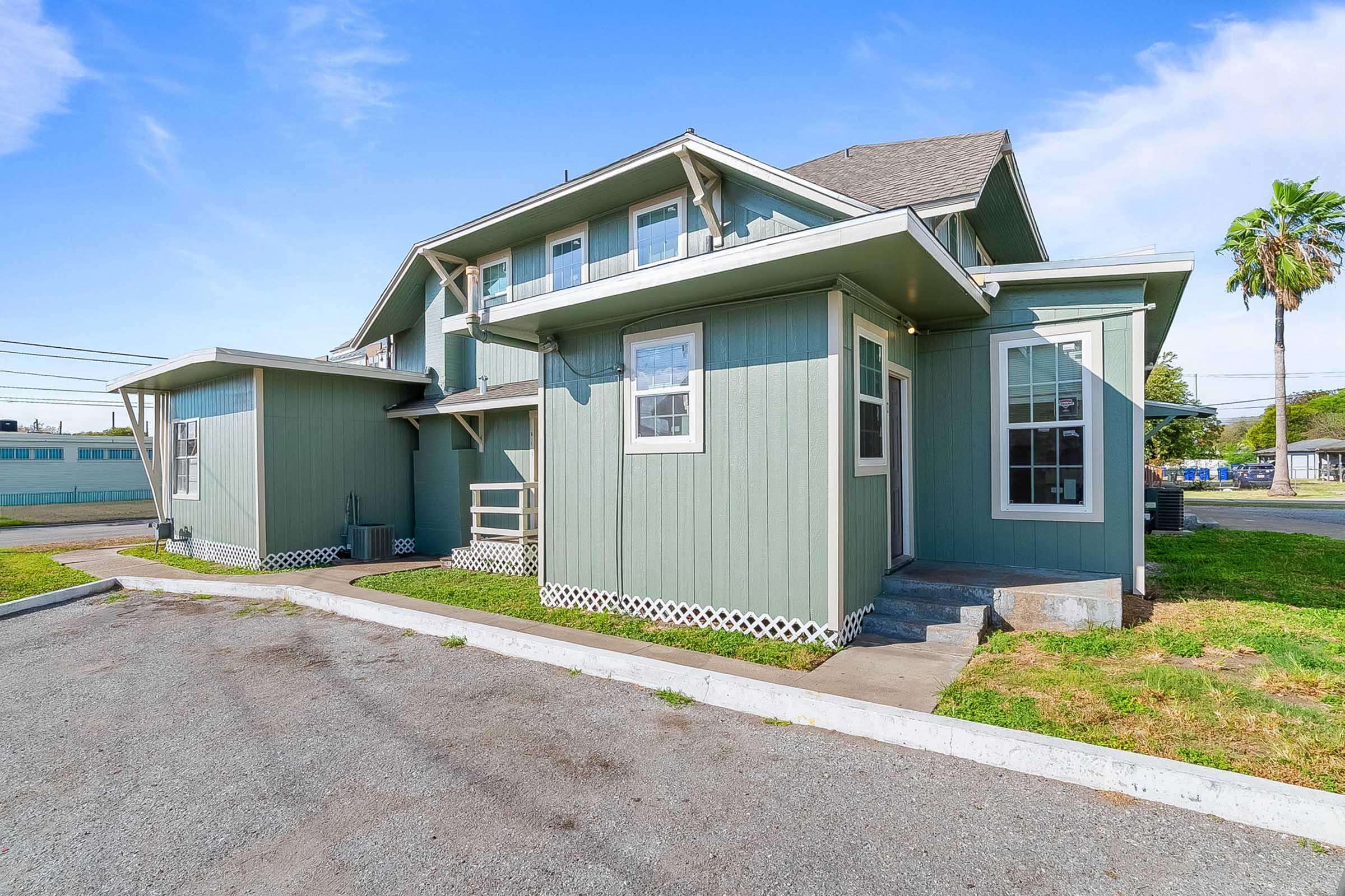 A two-story green house with a spacious porch and multiple windows. The front yard is grassy, and there are palm trees visible nearby. The building features a unique architectural design with a mix of angles and a shingled roof. The sky is clear with minimal clouds.