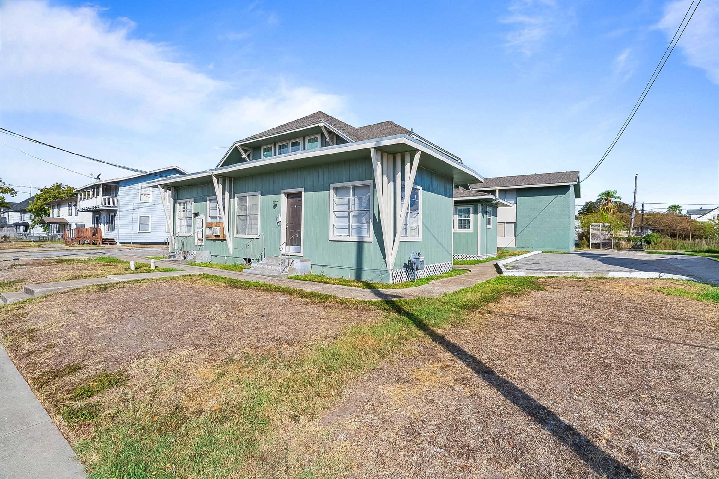 A light green multi-unit building with a flat roof, surrounded by dry grass and a concrete pathway. Power lines run overhead, and there are neighboring buildings in the background. The sky is blue with a few clouds.