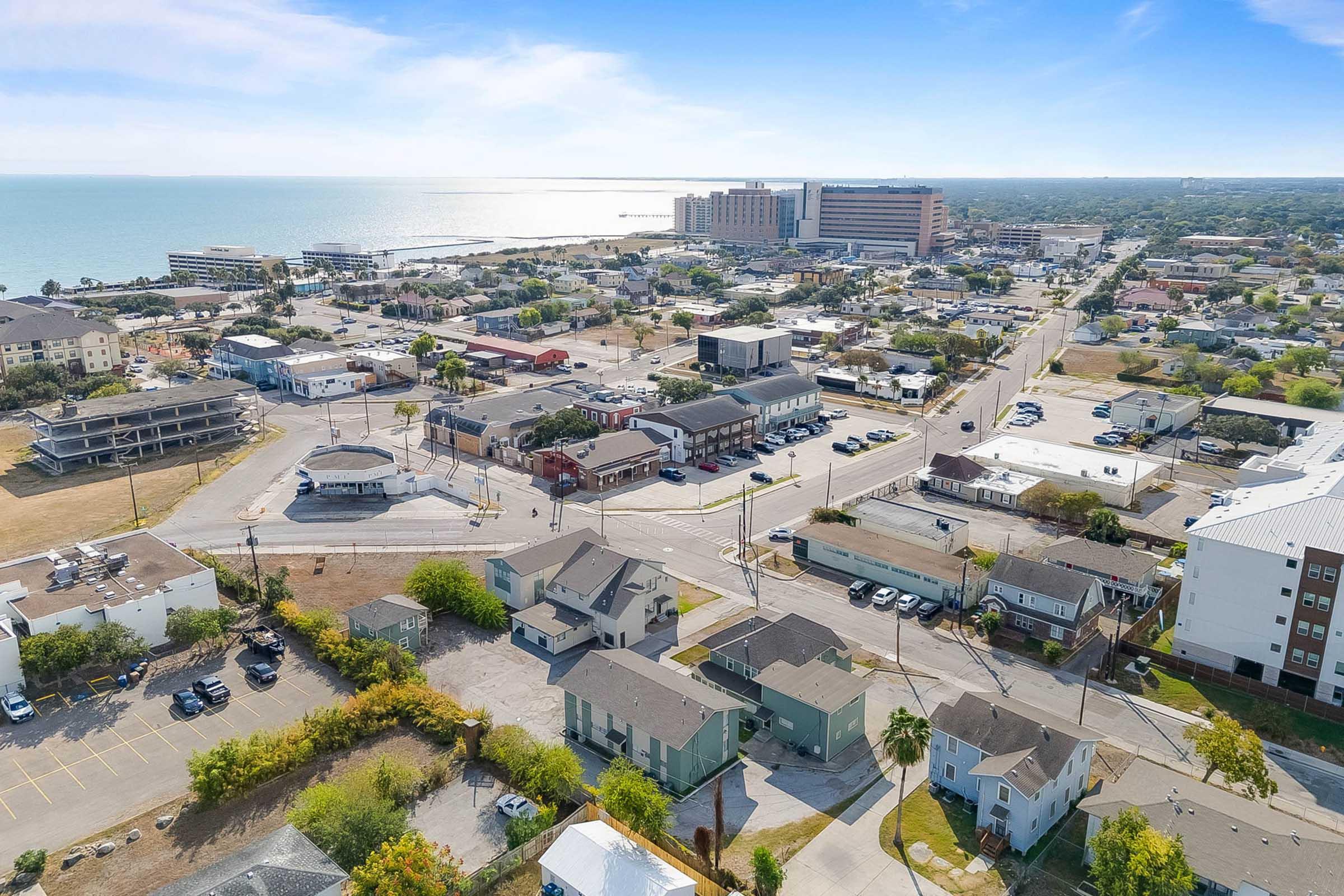 Aerial view of a coastal town with a mix of residential and commercial buildings. The scene includes a road intersection, parking lots, and proximity to the waterfront. In the background, a large building is visible, along with various smaller structures and green areas. The sky is clear and bright, suggesting a sunny day.