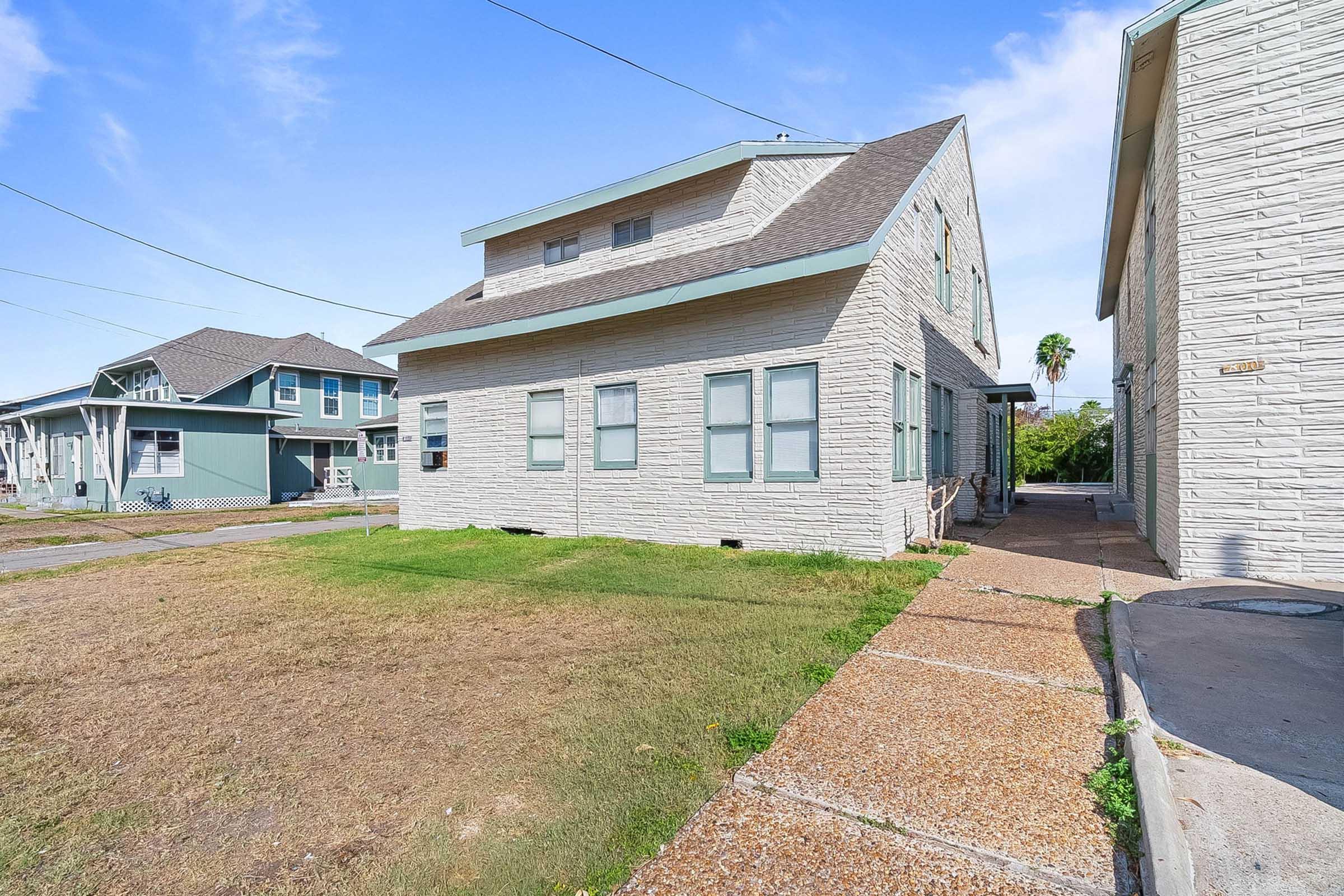 A two-story residential building with a light-colored exterior, featuring multiple windows and a gravel pathway leading to the entrance. The surrounding area includes a grassy lawn and additional houses in the background, set against a clear blue sky.