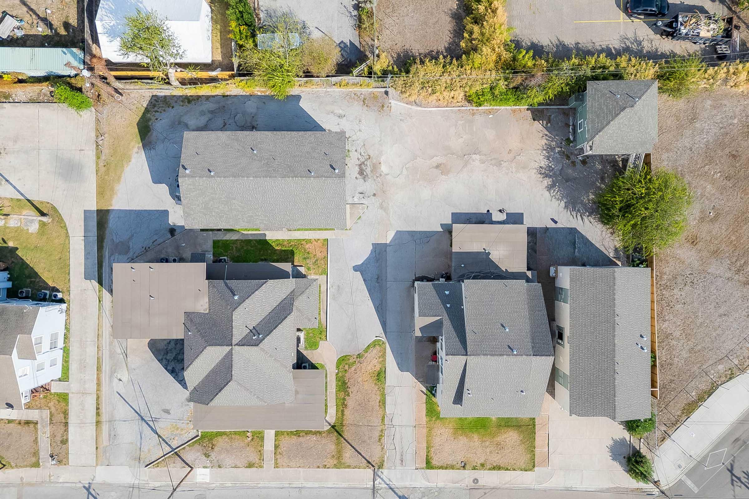 Aerial view of a residential area showing several houses with gray roofs, arranged in a grid layout. The surrounding ground is a mix of paved driveways and dirt areas, with a few trees visible. Some houses have yards, and there is a road running alongside the arrangement of homes.