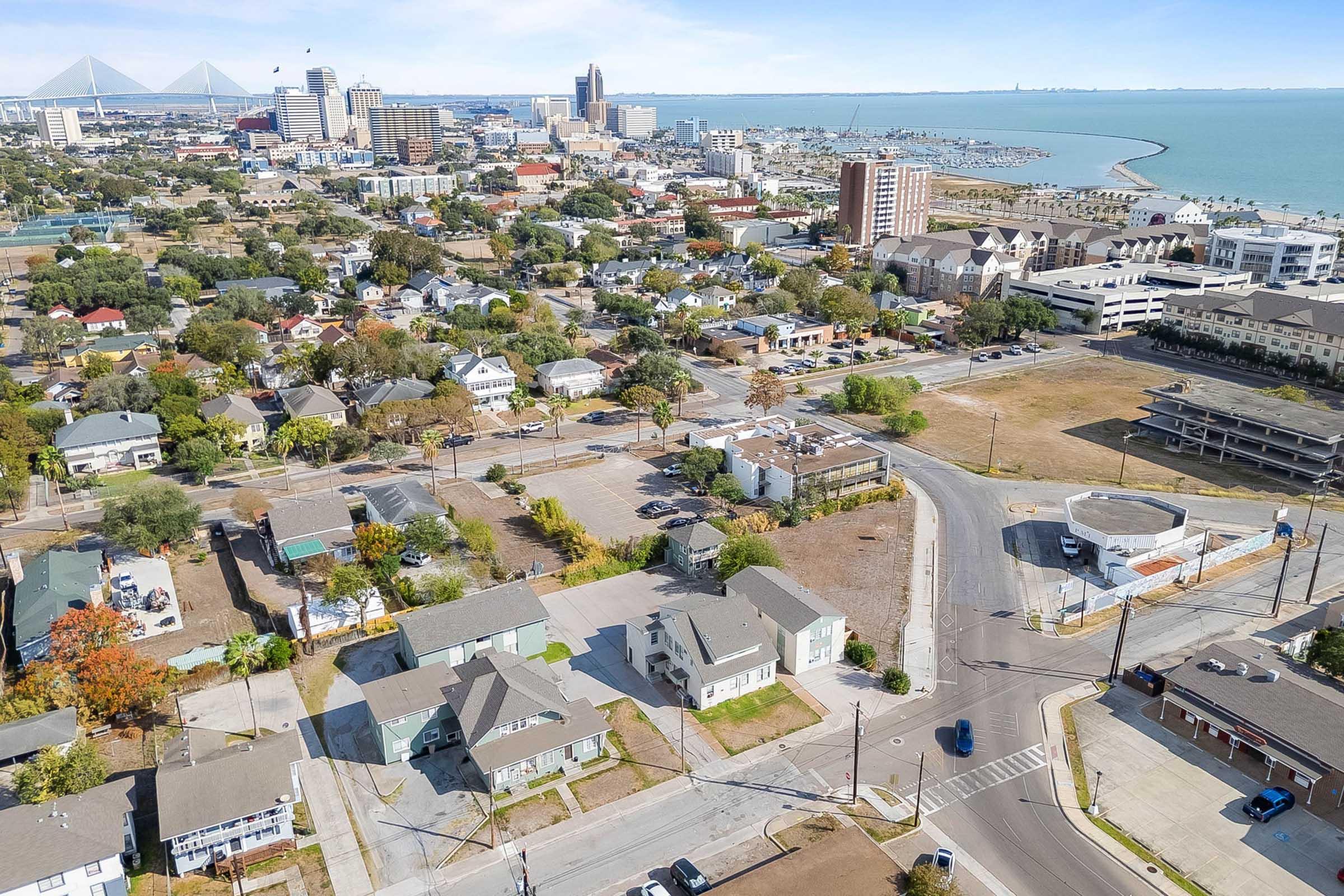 Aerial view of a coastal city featuring a mix of residential homes, hotels, and commercial buildings, with the ocean and harbor visible in the background. The skyline includes various high-rise structures, alongside roads and greenery, showcasing urban development near the waterfront.