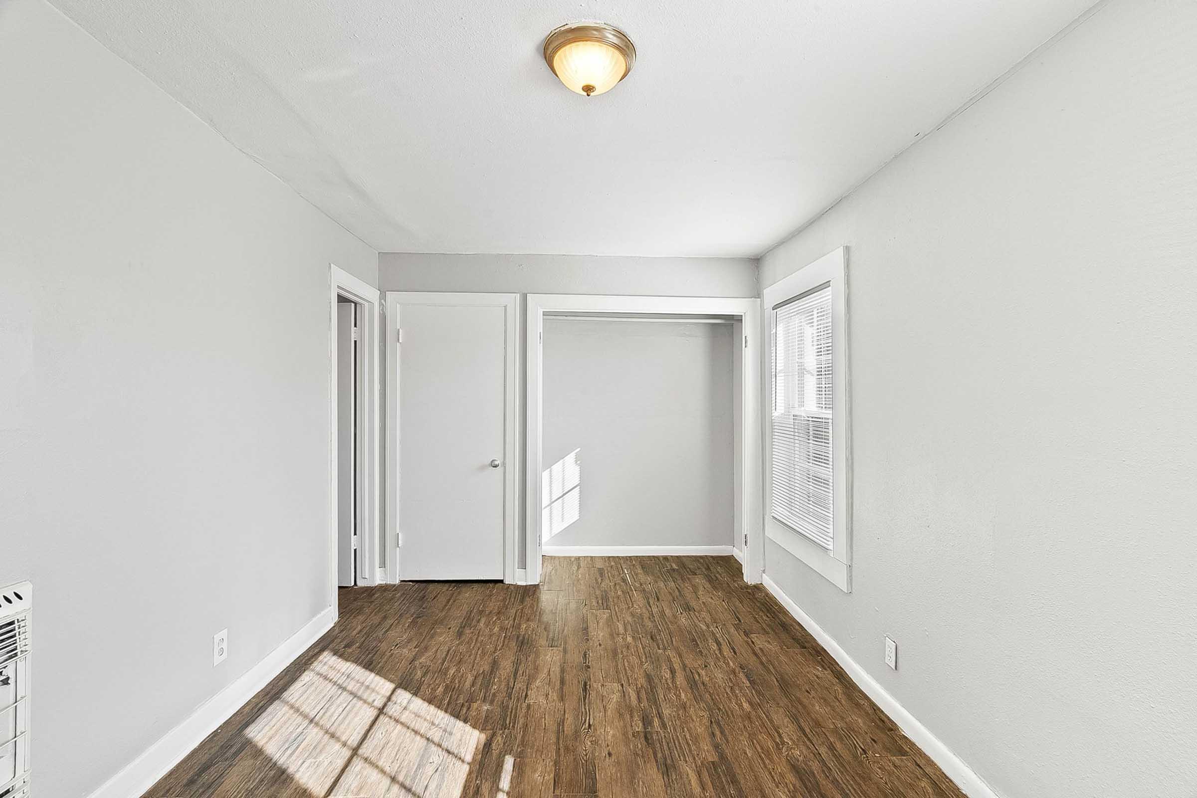 A well-lit, empty room with gray walls and hardwood flooring. It features a ceiling light, a closet with a white door, and a large window allowing natural light to stream in, casting shadows on the floor. There are two additional doors leading to other spaces, enhancing the room's openness.