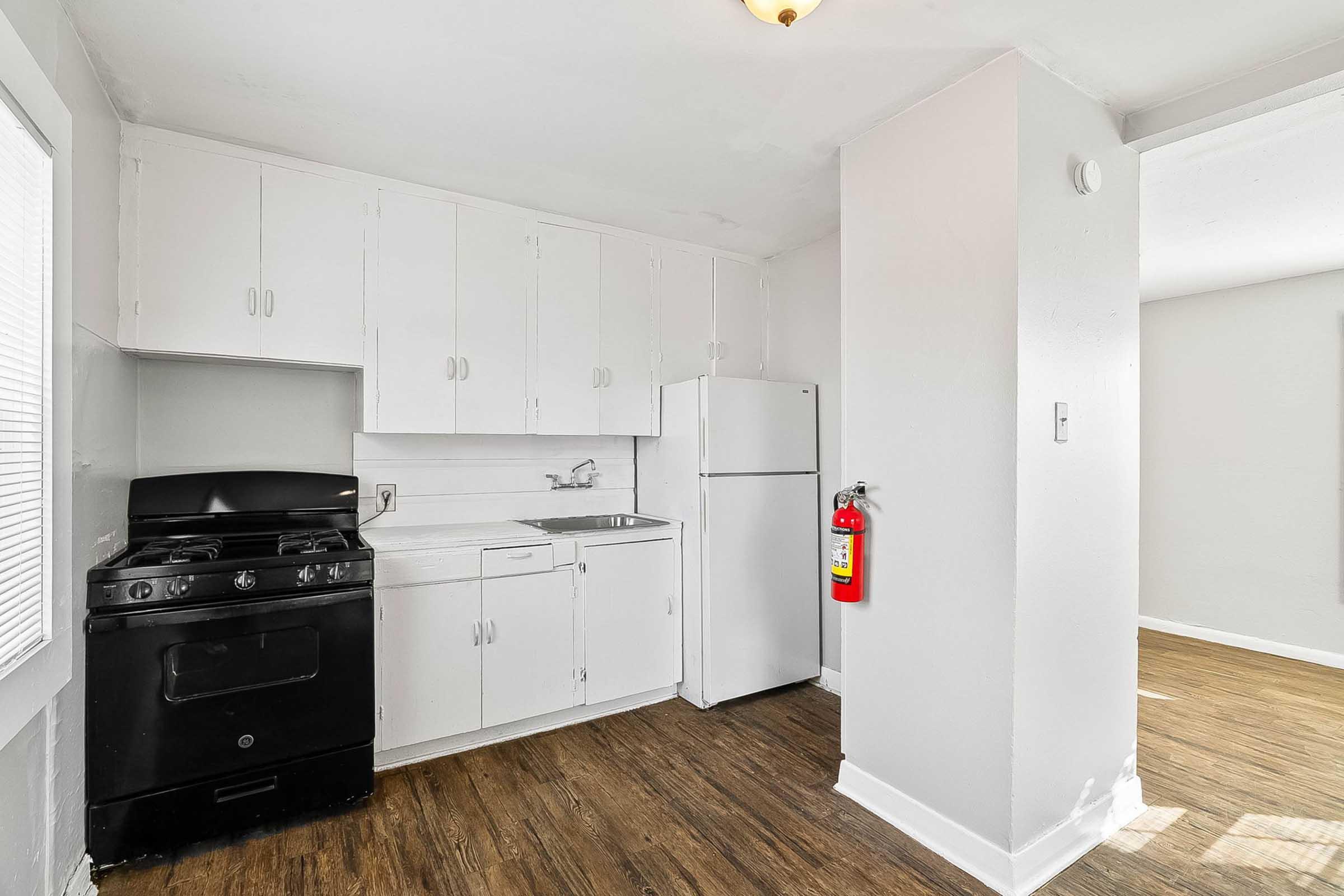 A small, minimalist kitchen featuring white cabinets, a black gas stove, a white refrigerator, and a sink. The flooring is wooden, and a fire extinguisher is mounted on the wall. Natural light enters through a window.