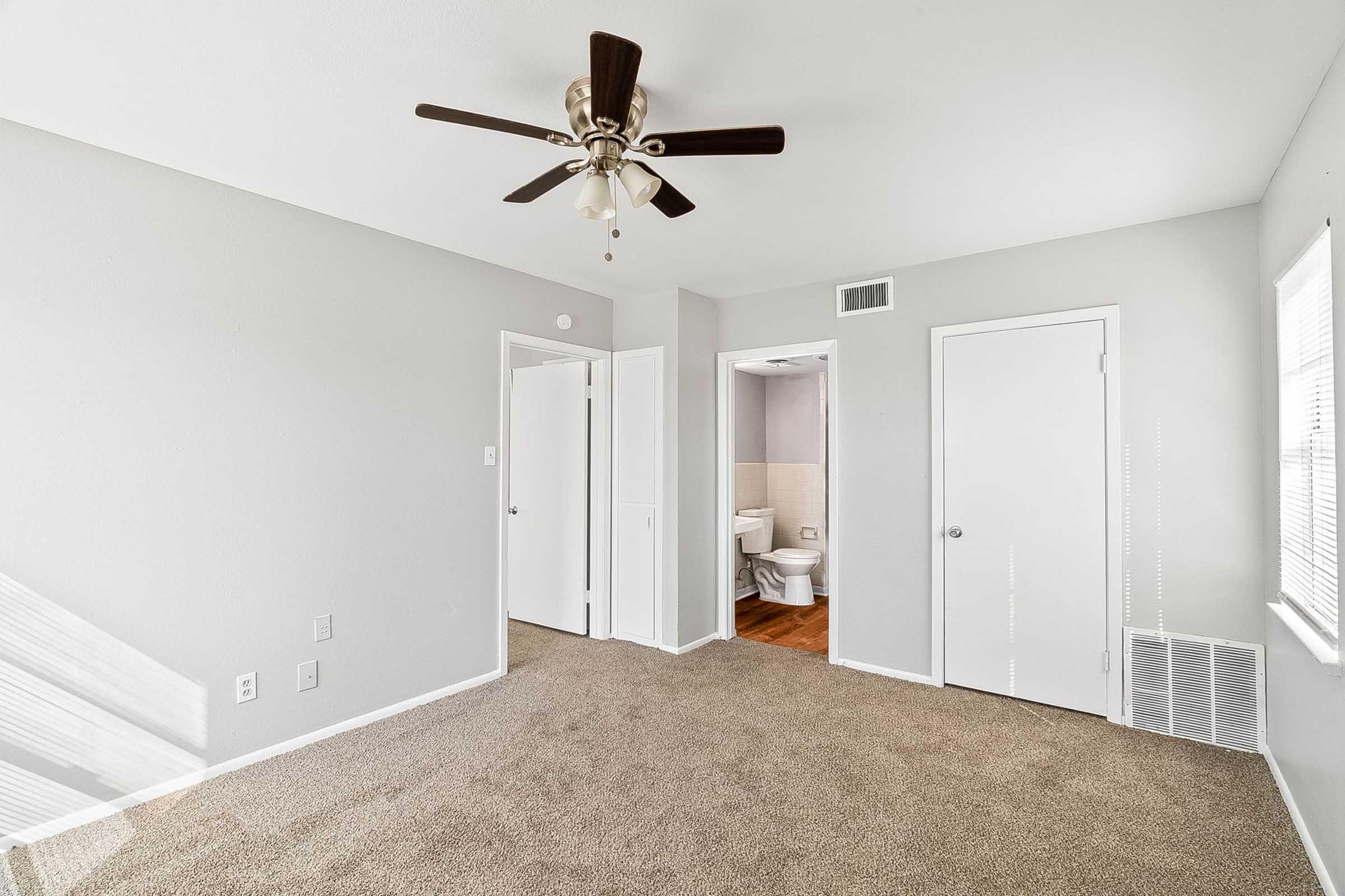 A spacious, light-filled bedroom featuring beige carpet, a ceiling fan with wooden blades, and neutral gray walls. There are two closed doors leading to other areas of the home, and a glimpse of a bathroom is visible through one of the doors. Natural light comes from a nearby window.