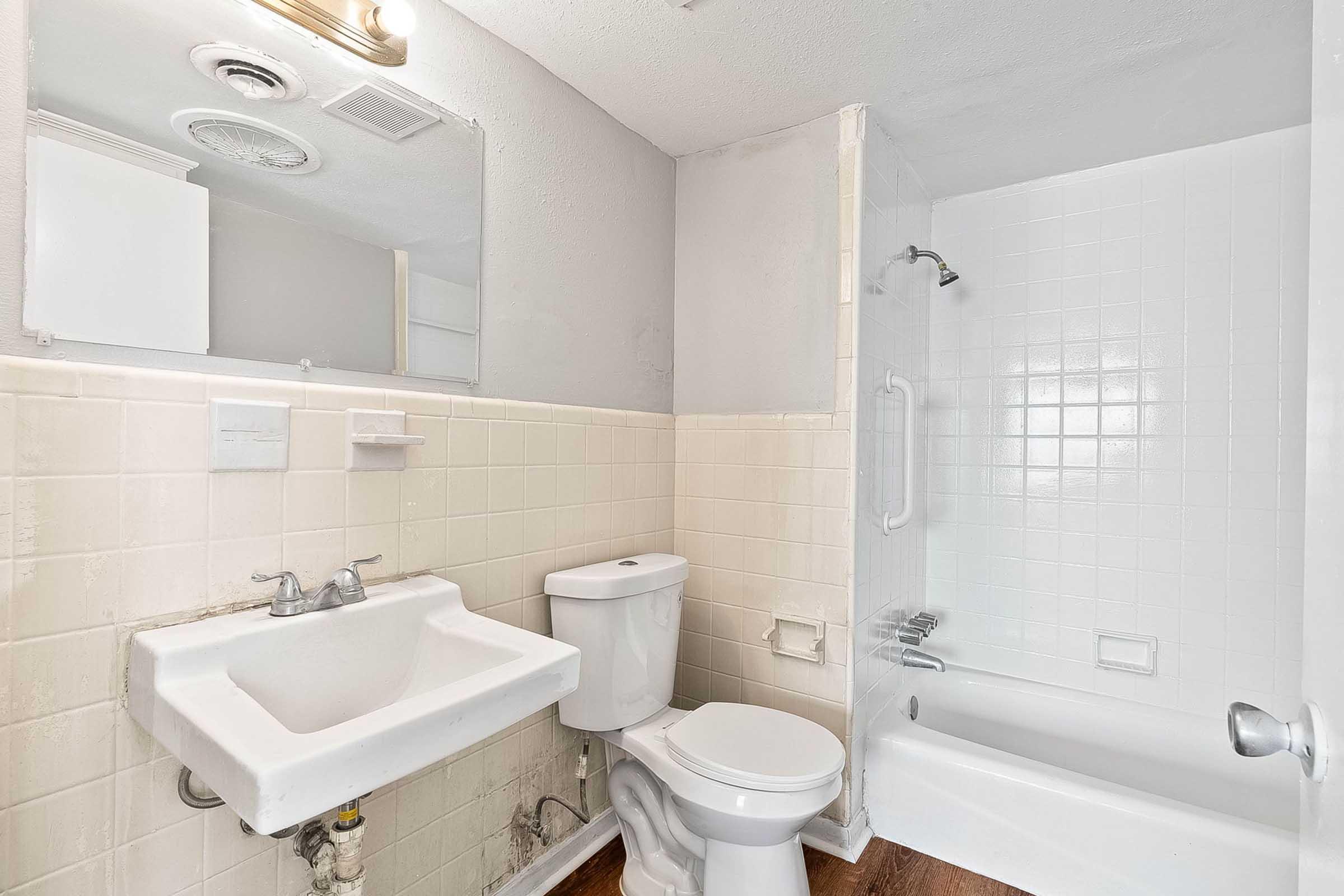 A small bathroom featuring a white sink, toilet, and bathtub with shower. The walls are partially tiled in light colors, and there's a mirror above the sink. The floor appears to be wood-like.