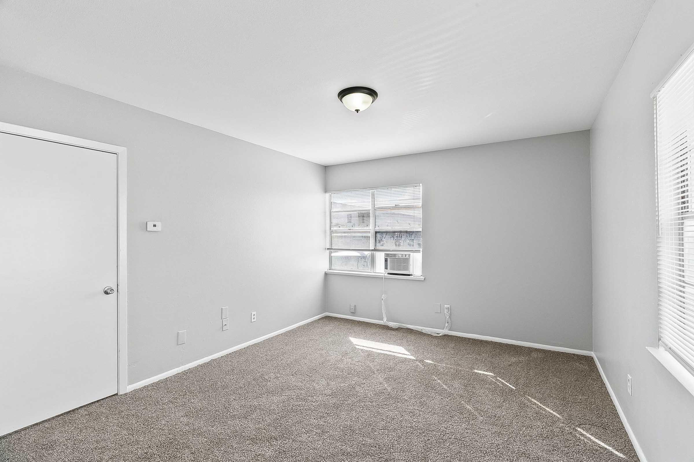 A lightly furnished room with gray walls and carpet. It features a single window allowing natural light, a ceiling light fixture, and a white door. There are no visible decorations or furniture, creating a spacious and minimalistic atmosphere.