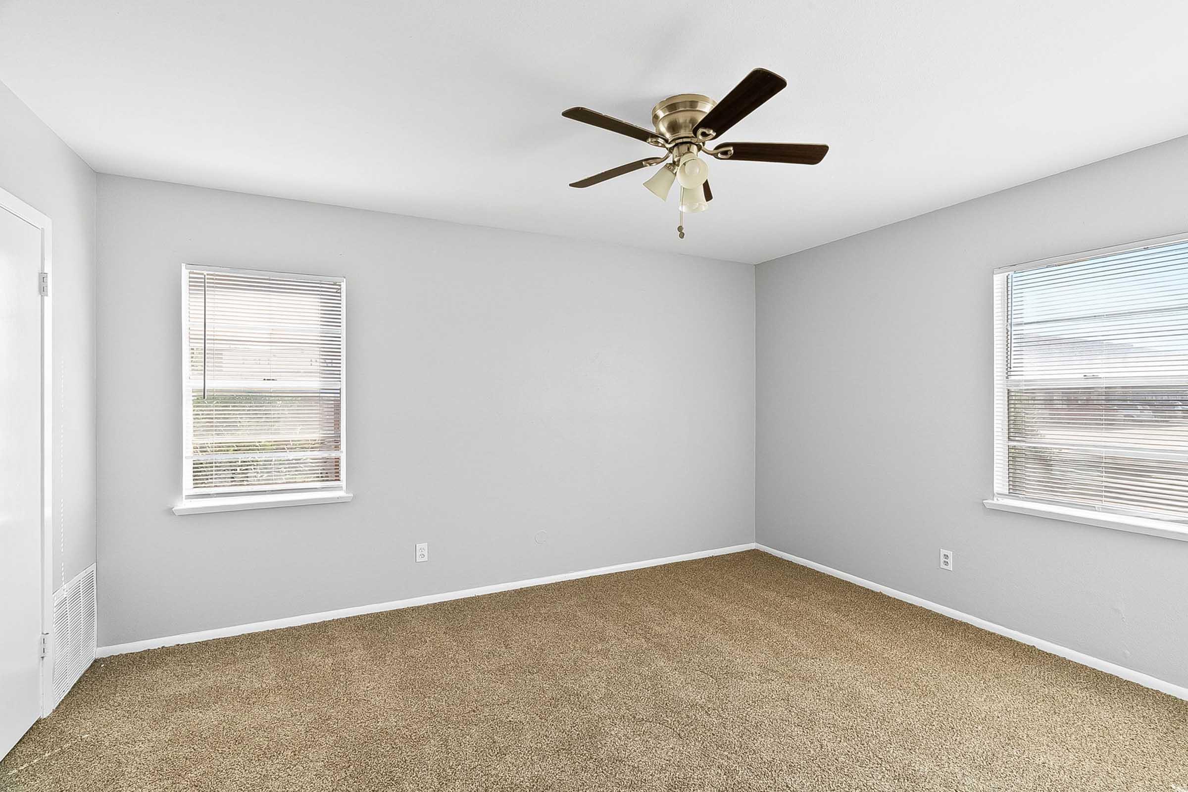 A clean, empty room with light gray walls and two windows with blinds. The floor is covered with beige carpet, and a ceiling fan is mounted in the center of the room, adding a warm and inviting touch. Natural light illuminates the space, creating a bright atmosphere.