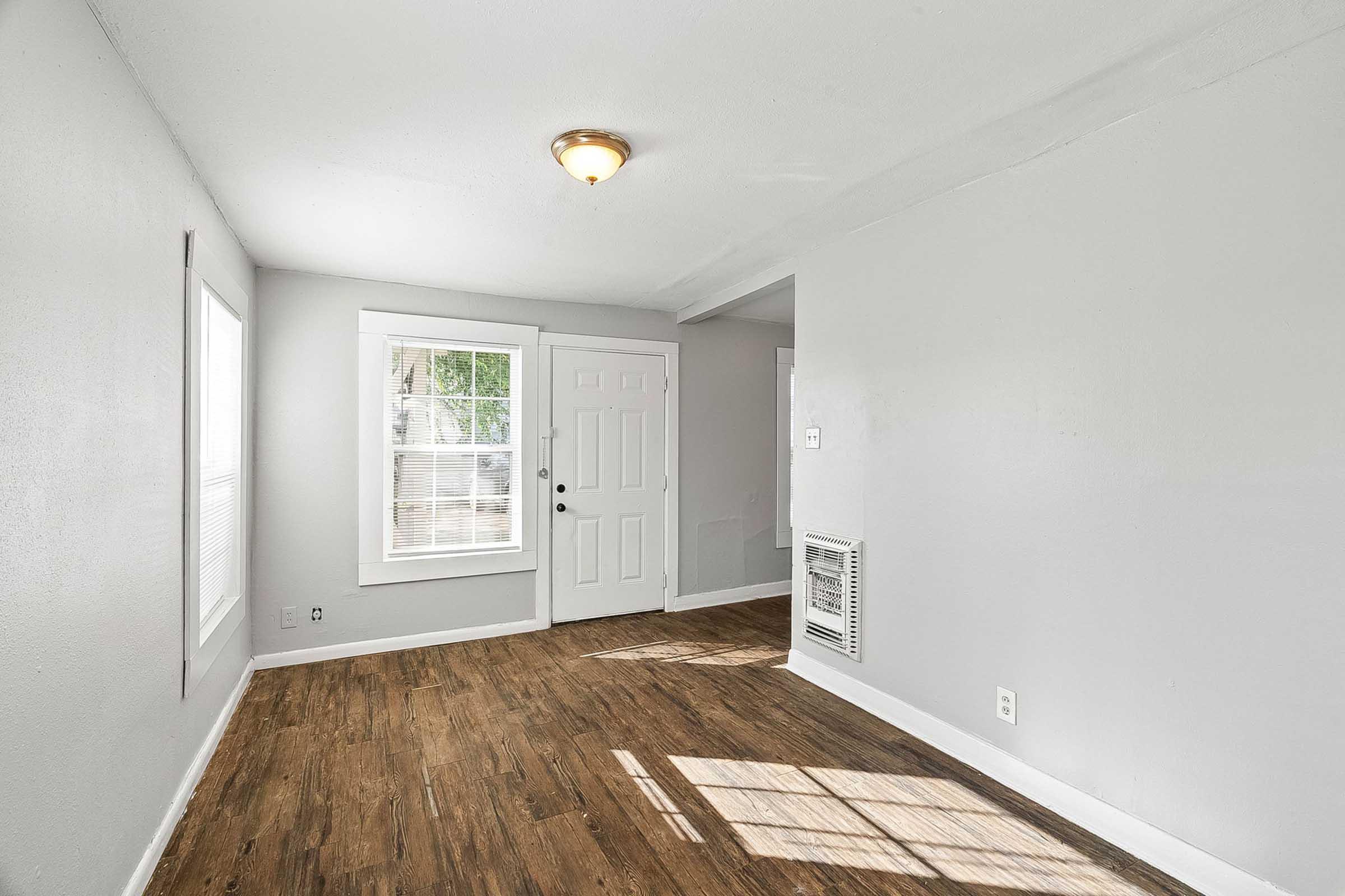 A bright, empty room with light gray walls and hardwood flooring. There is a white door leading outside, two windows allowing natural light in, and a wall-mounted heater. The space appears clean and well-maintained, suitable for various uses.