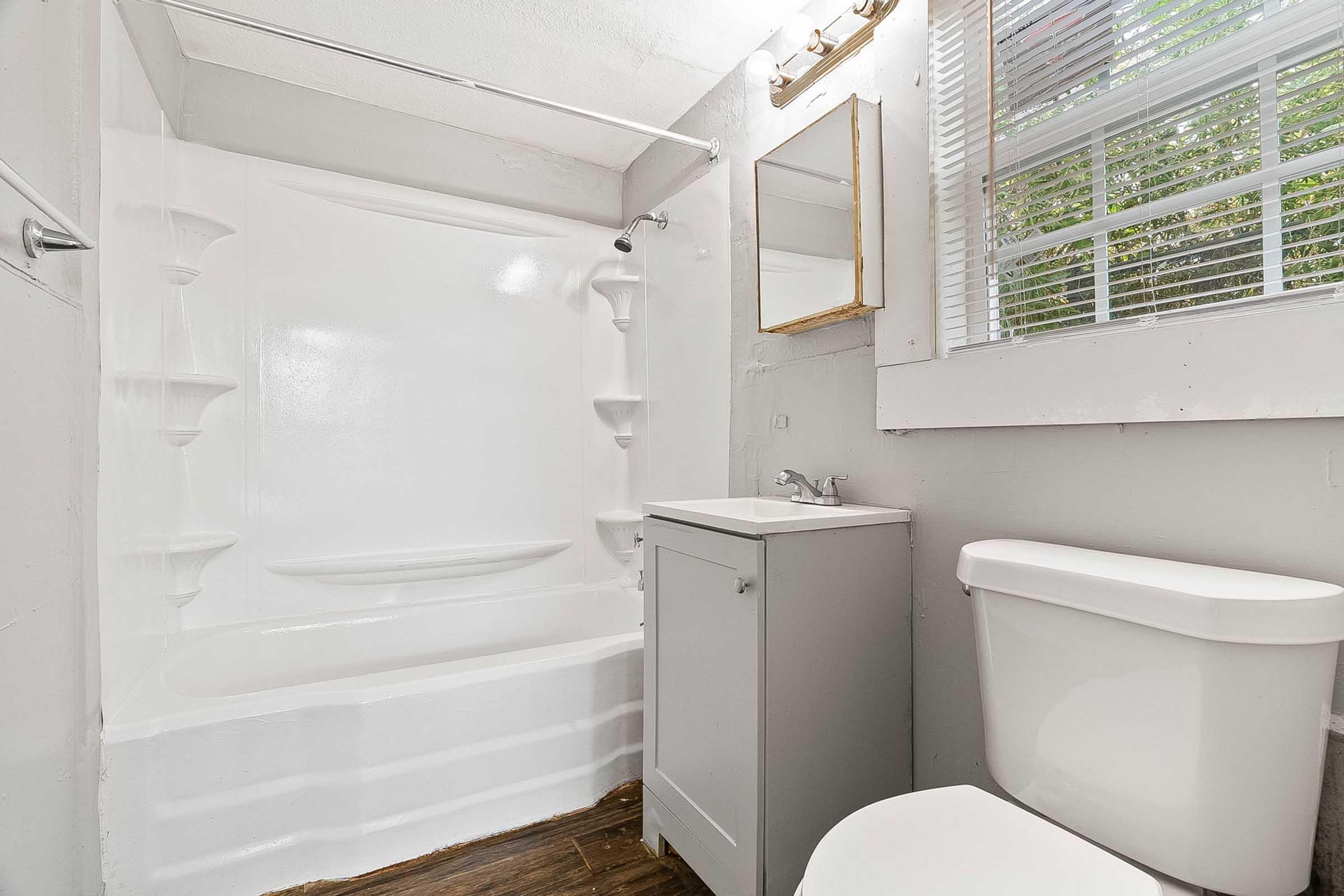 A clean, modern bathroom featuring a white bathtub with integrated shelves, a compact sink cabinet, a white toilet, and a window with blinds allowing natural light. The walls are painted light gray, and there’s a mirror above the sink. The flooring is dark wood, creating a contrasting aesthetic.
