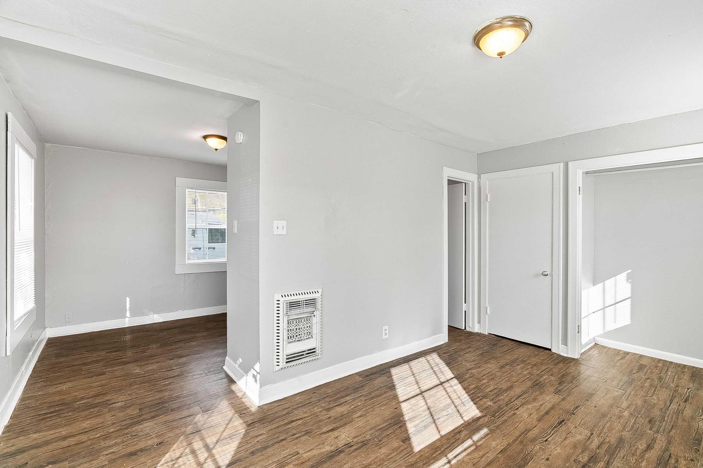 A bright, airy interior of a room featuring light gray walls and hardwood flooring. There is a wall-mounted heater visible, and two doorways lead to other areas. Natural light streams in through a window, creating a welcoming atmosphere. The room is empty and uncluttered, suggesting a fresh living space.