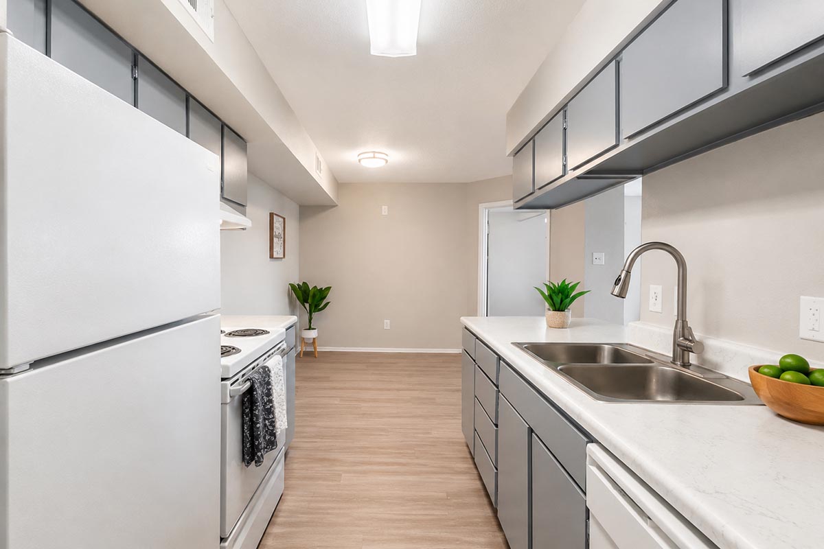 A modern kitchen featuring white cabinets, a stainless steel sink, and a stove. The countertops are light-colored with decorative plants placed on them. The kitchen opens up to a spacious living area with neutral walls and flooring, illuminated by overhead lighting.