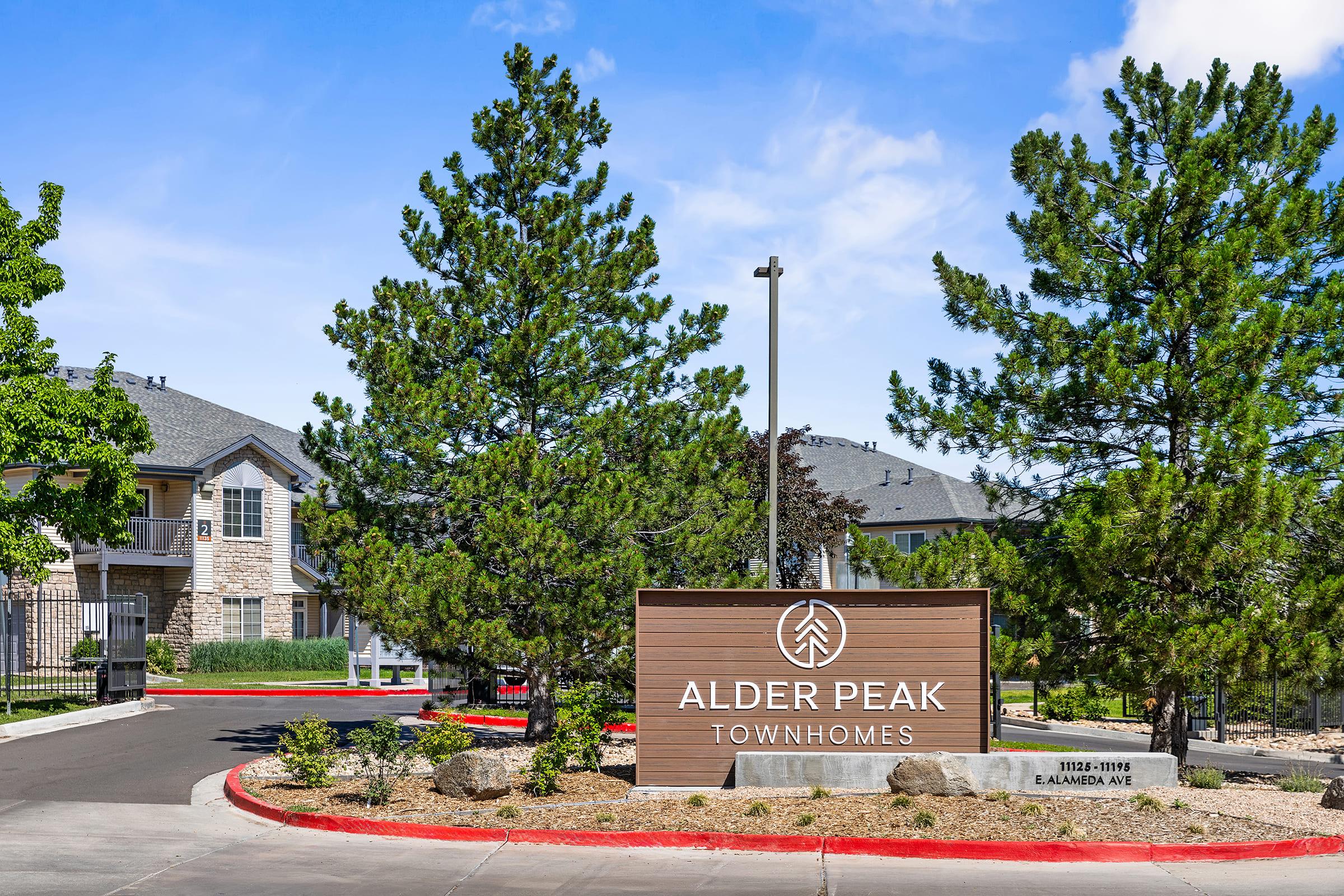 Signage for Alder Peak Townhomes, featuring a wooden structure with the name prominently displayed. Surrounding the sign are landscaped areas with rocks and small trees under a clear blue sky. The entrance is paved with a red-bordered driveway leading into the complex.