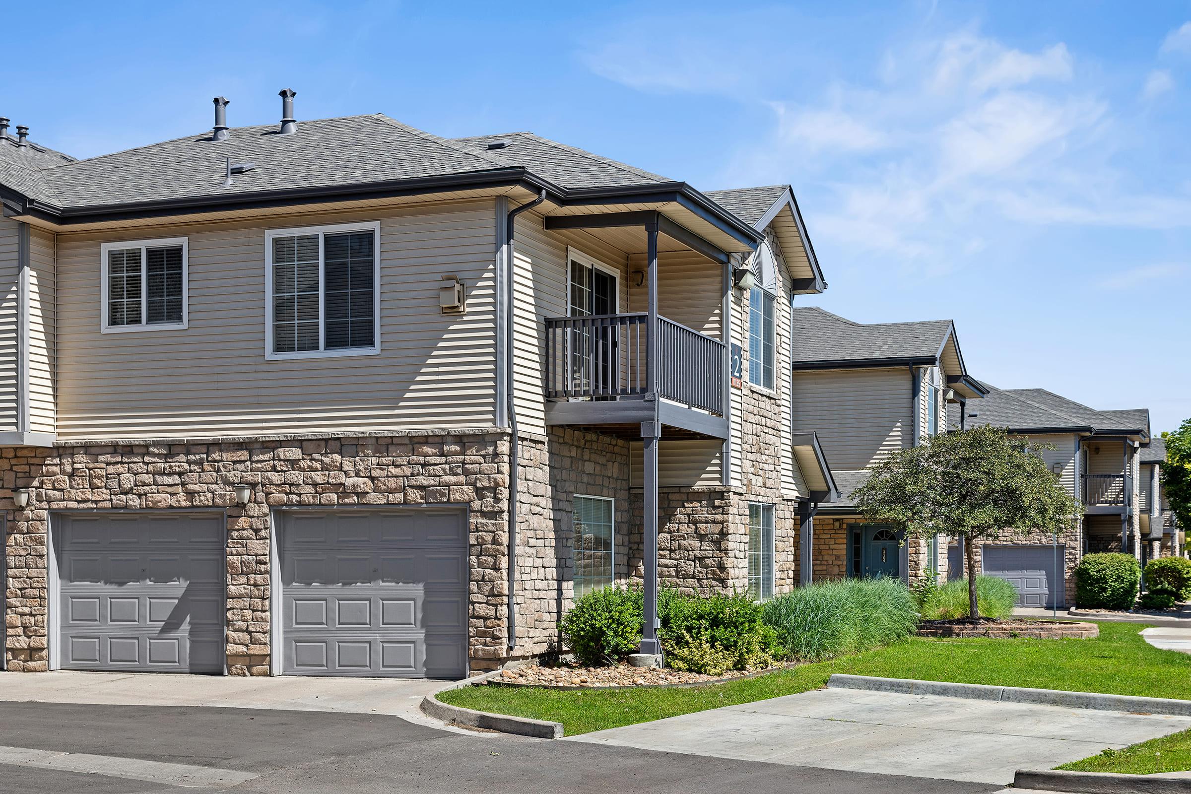 A view of a modern residential building featuring a blend of beige siding and stone facade. The structure has a balcony and garage doors on the ground floor. Surrounding greenery, a paved driveway, and clear blue skies create a welcoming suburban atmosphere.