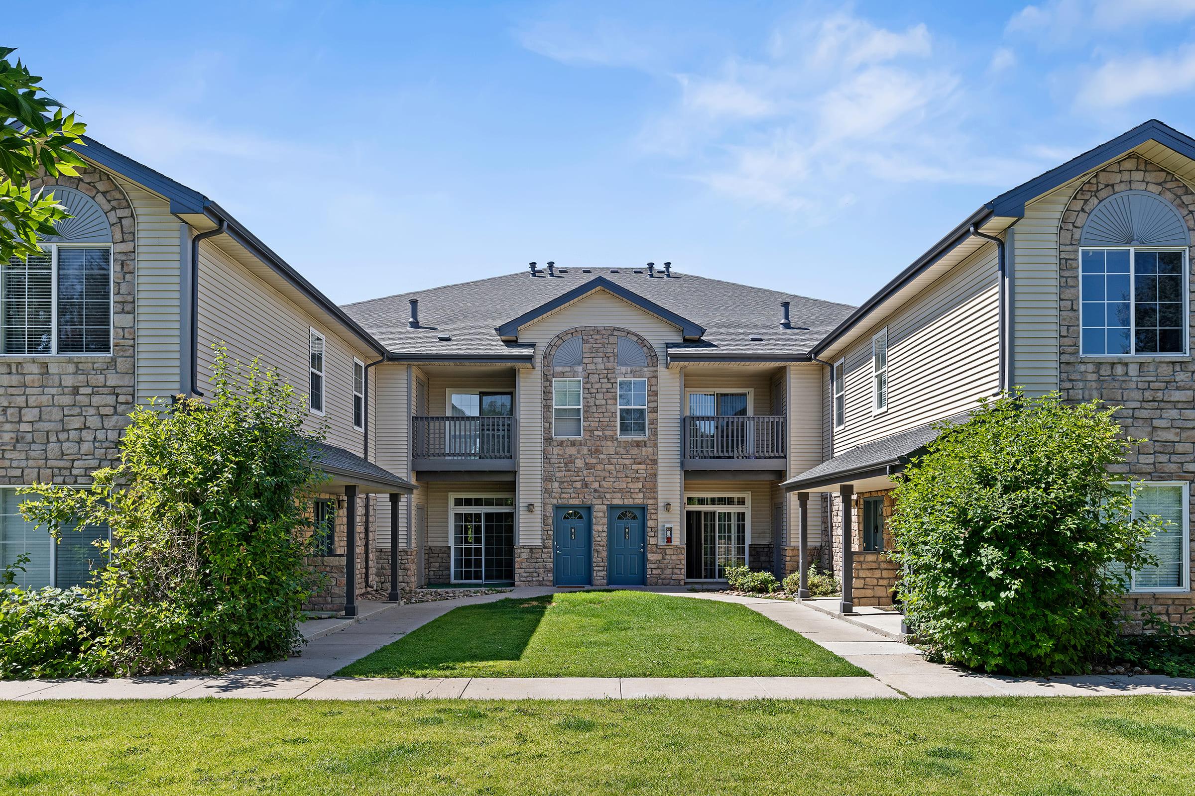 Two-story apartment building with stone and siding exterior. It features a central entrance flanked by small balconies on the upper floor, surrounded by lush green grass and landscaping. The clear blue sky provides a bright backdrop for the residential setting.