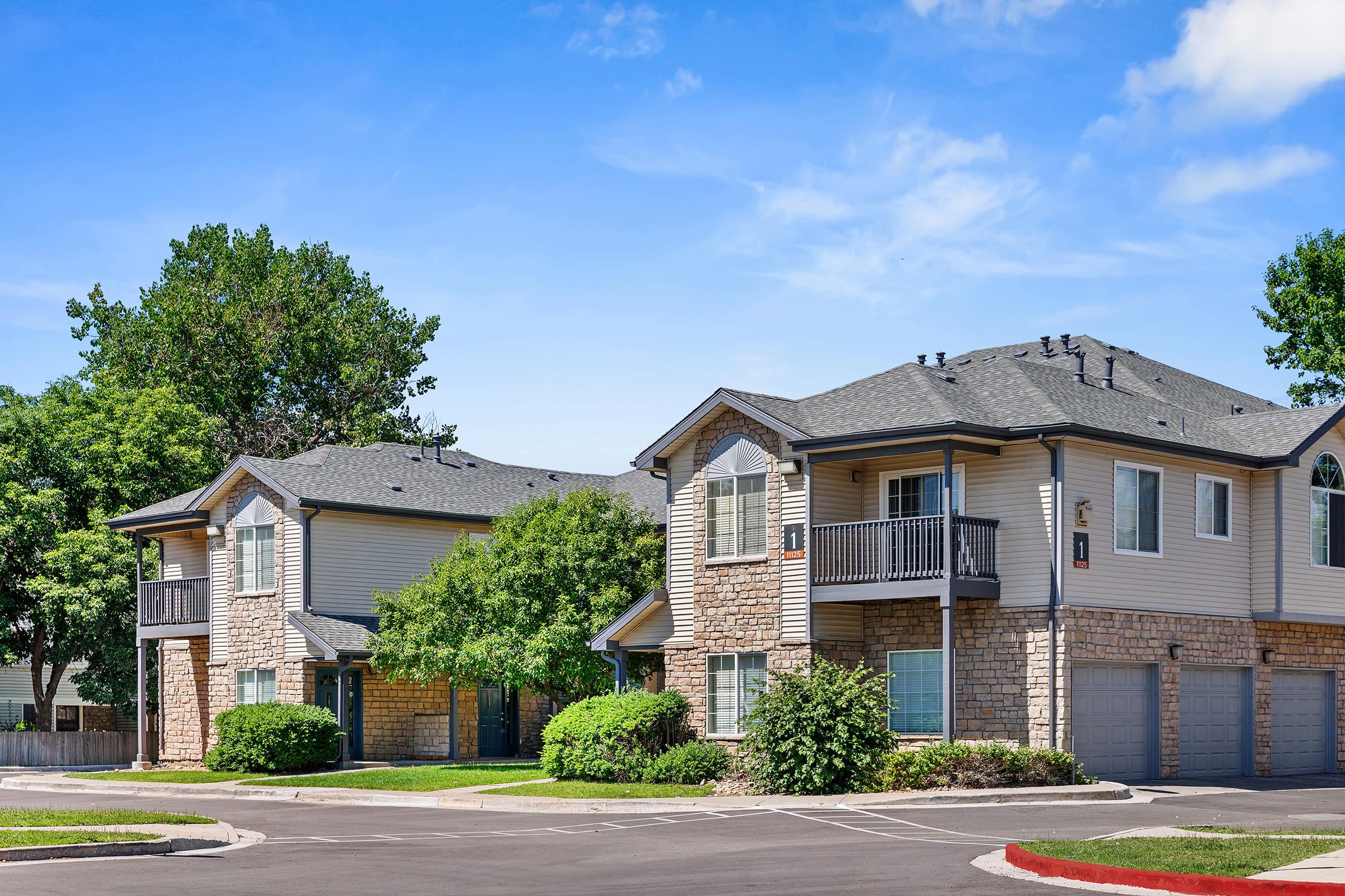 Two residential buildings with a mix of stone and siding exteriors, surrounded by lush green trees and shrubs. The sky is clear blue, and the setting appears clean and well-maintained. Parking spaces are visible in front of the homes.