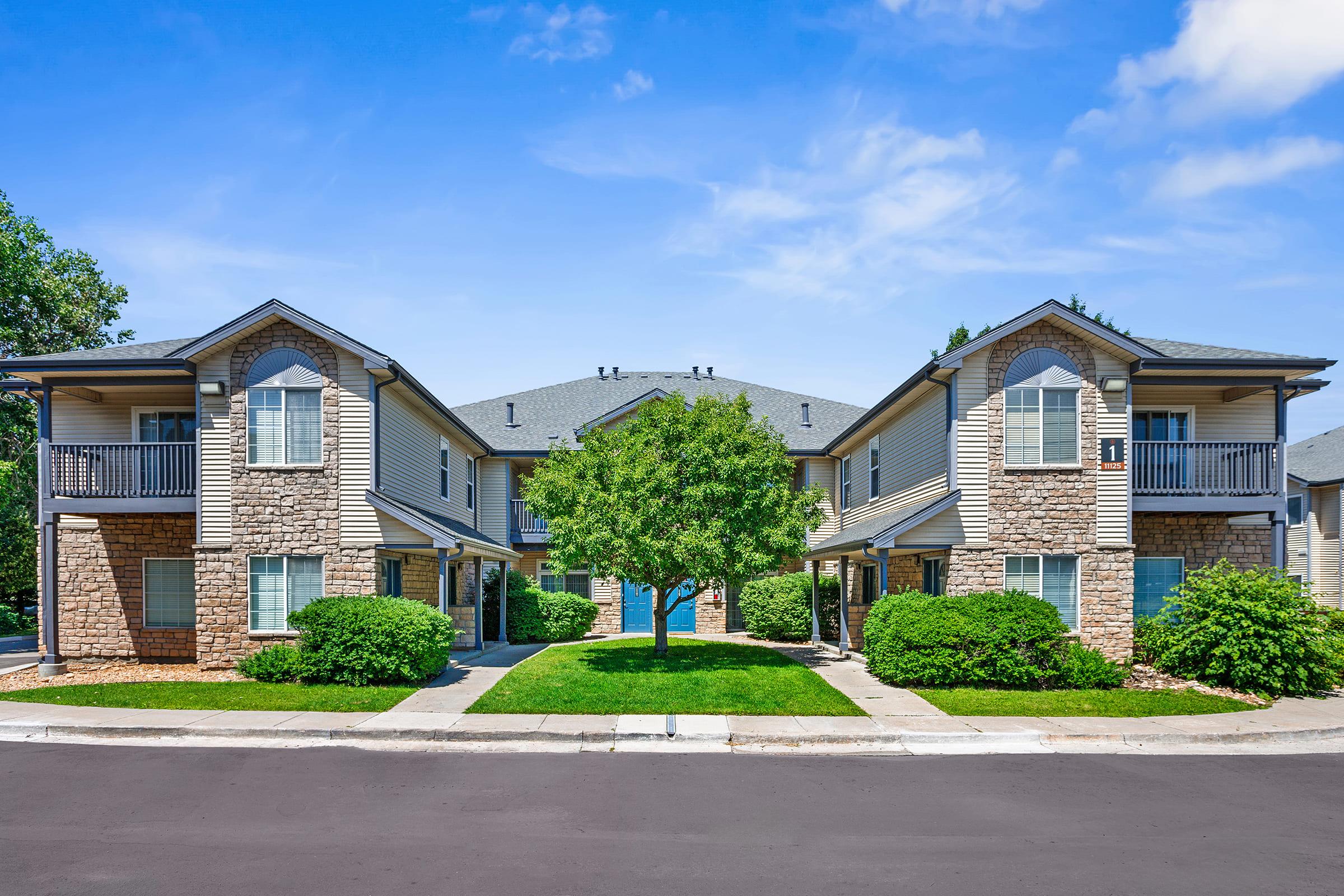 Two modern apartment buildings with stone accents, situated symmetrically around a green lawn and a small tree. The sky is clear and blue, and the well-maintained landscaping includes shrubs and a walkway leading to the apartments. The scene conveys a peaceful suburban atmosphere.