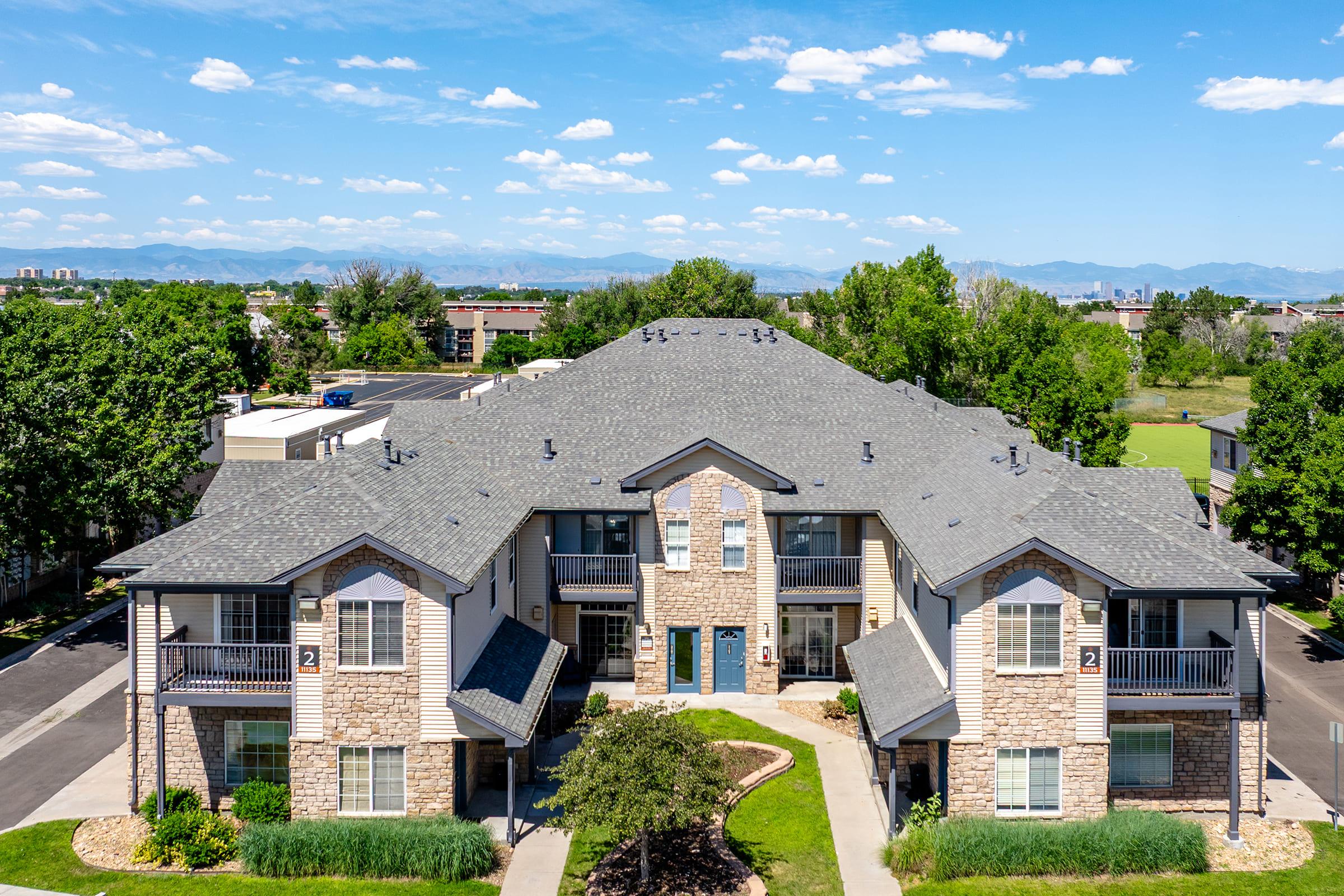 Aerial view of a multi-unit residential building with a stone facade, surrounded by green landscaping and trees. The sky is partly cloudy, and mountains are visible in the background. The building features a central entrance and is situated in a well-maintained area with parking spaces nearby.