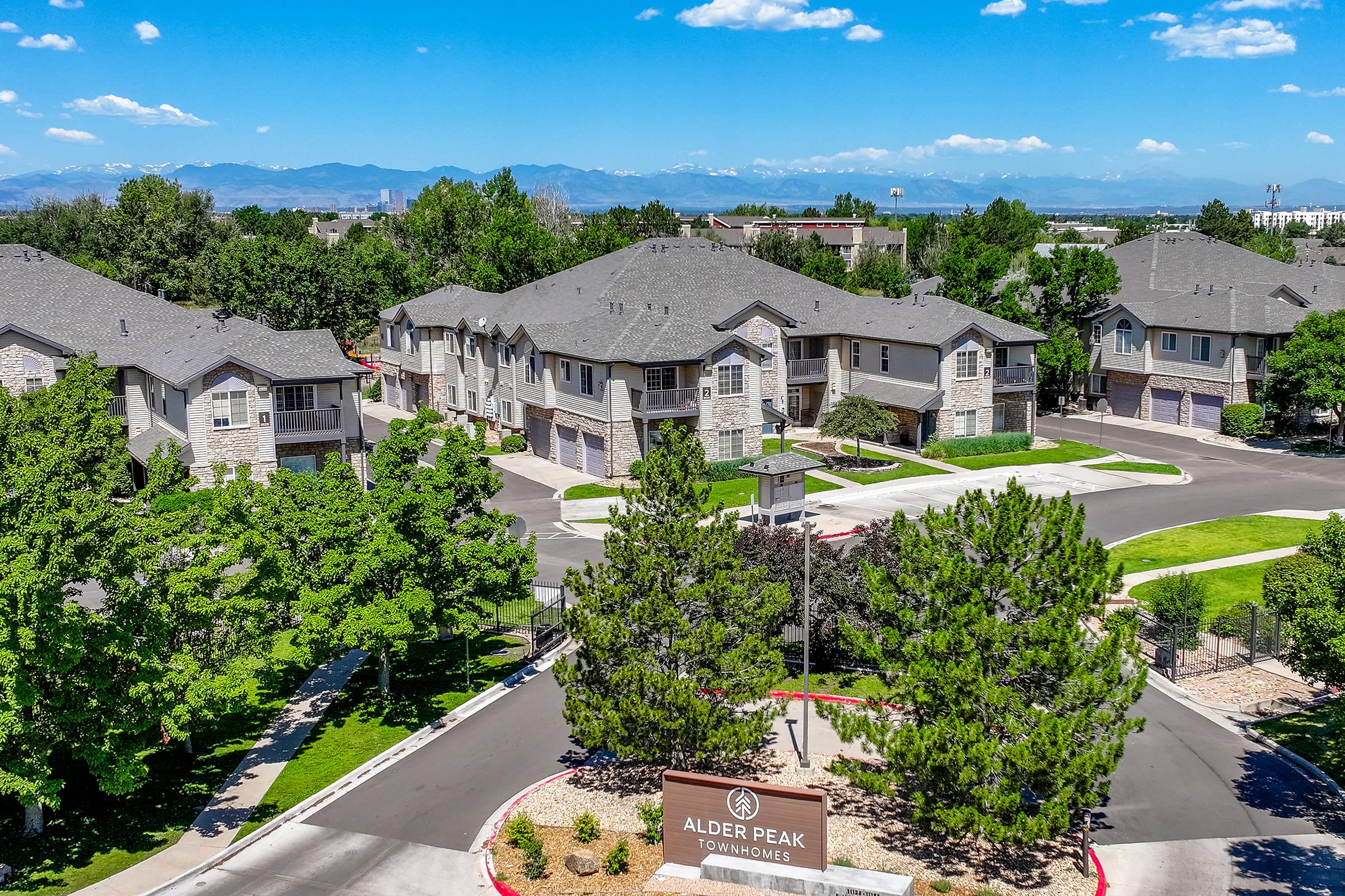 Alder Peak Townhomes, featuring multiple residential buildings surrounded by trees and landscaping, with a clear blue sky and distant mountains in the background. The scene includes a welcome sign and well-maintained roads, showcasing a peaceful suburban environment.