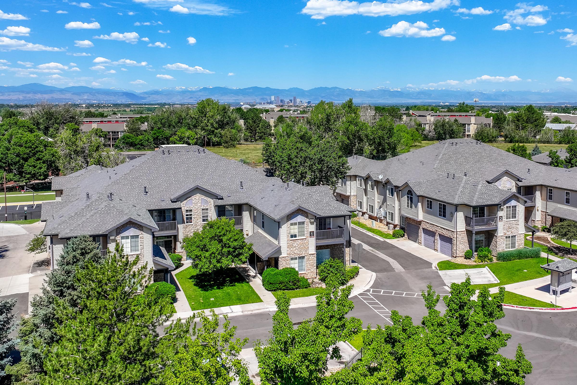 Aerial view of a residential community featuring multiple buildings with a mix of stone and siding exteriors. Lush green trees and well-maintained landscaping surround the grounds. In the background, rolling hills and mountains are visible under a blue sky with fluffy white clouds.