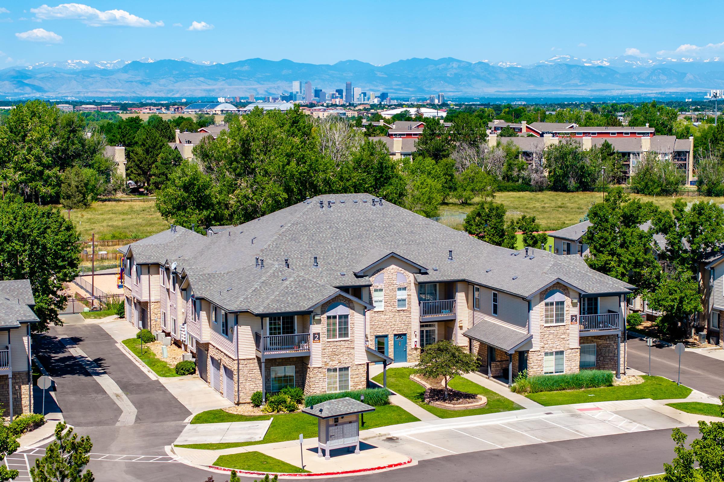 A residential neighborhood featuring several modern apartment buildings with a mix of stone and siding exteriors. Lush greenery surrounds the area, and the distant skyline of a city can be seen beneath a clear blue sky with mountains in the background, creating a picturesque urban-suburban landscape.