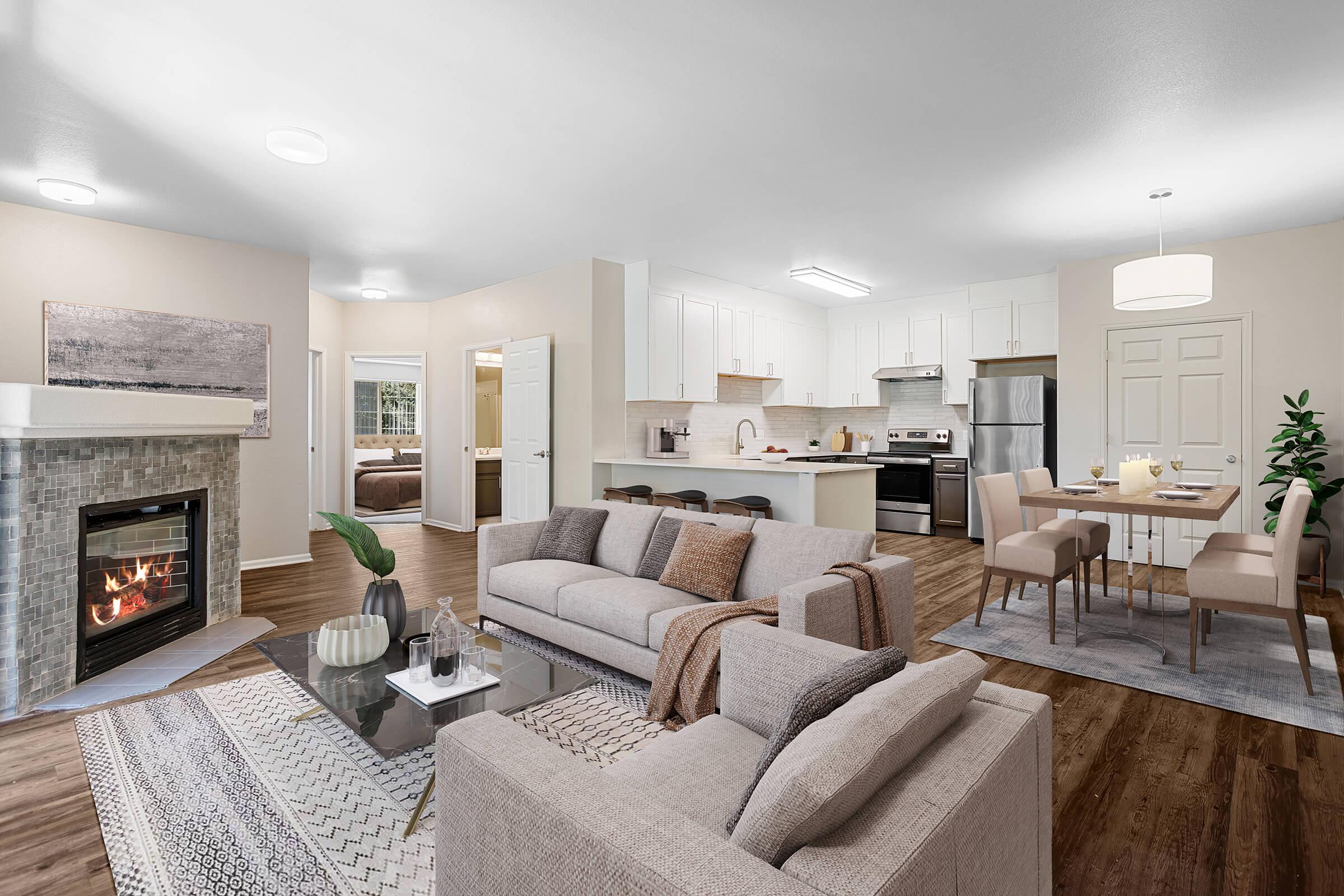 Modern open-concept living space featuring a cozy seating area with a fireplace, a dining area with a table set for four, and a well-equipped kitchen. Soft neutral colors and warm wood flooring create a welcoming atmosphere. Large windows allow natural light to flow in, enhancing the inviting ambiance.