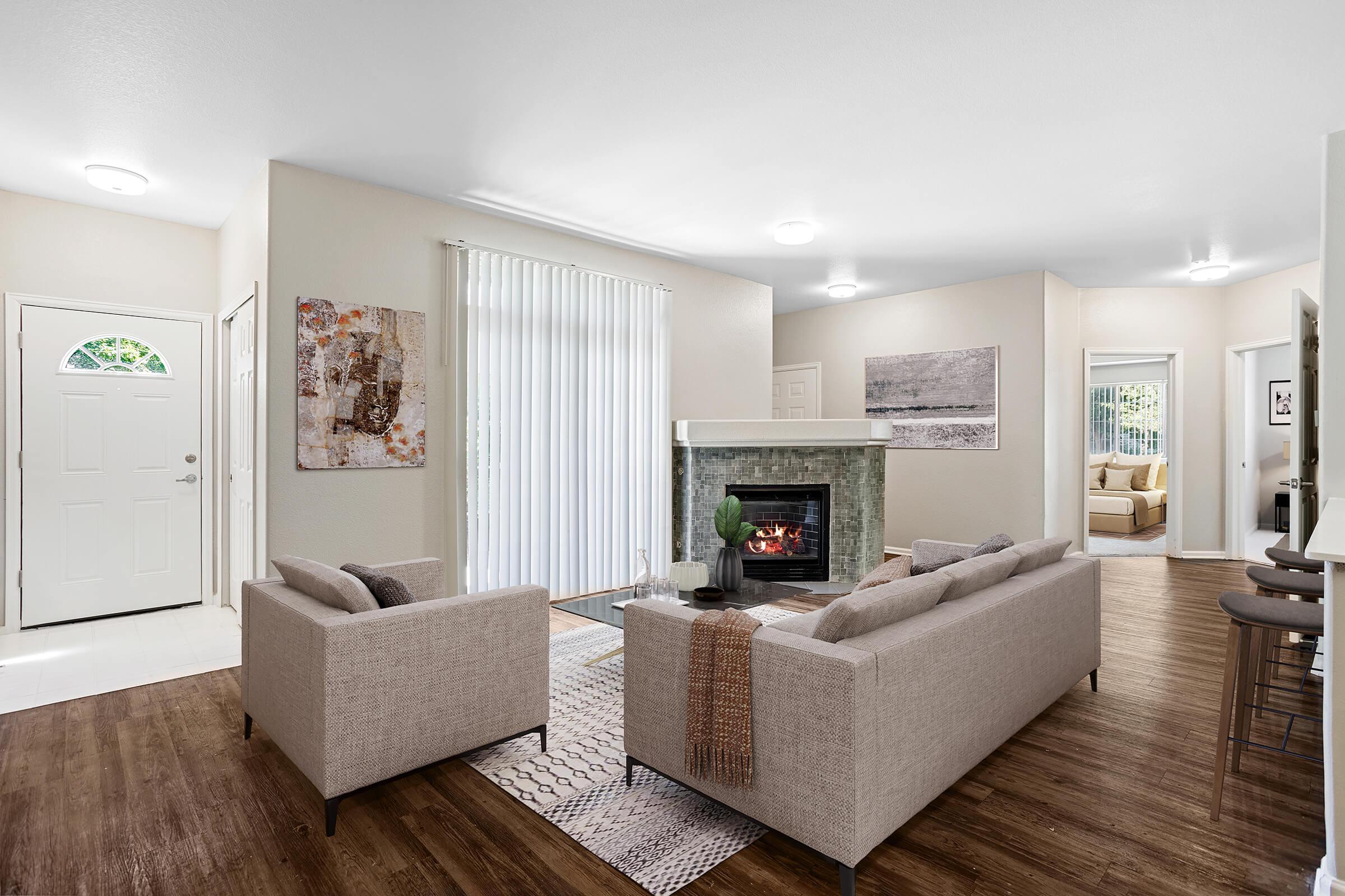 Bright and spacious living room featuring two light-colored sofas facing a modern fireplace. Large windows with vertical blinds allow natural light in. A doorway leads to a welcoming entrance, while a neutral color palette creates a cozy atmosphere. Decorative wall art and a rug add warmth and style to the space.