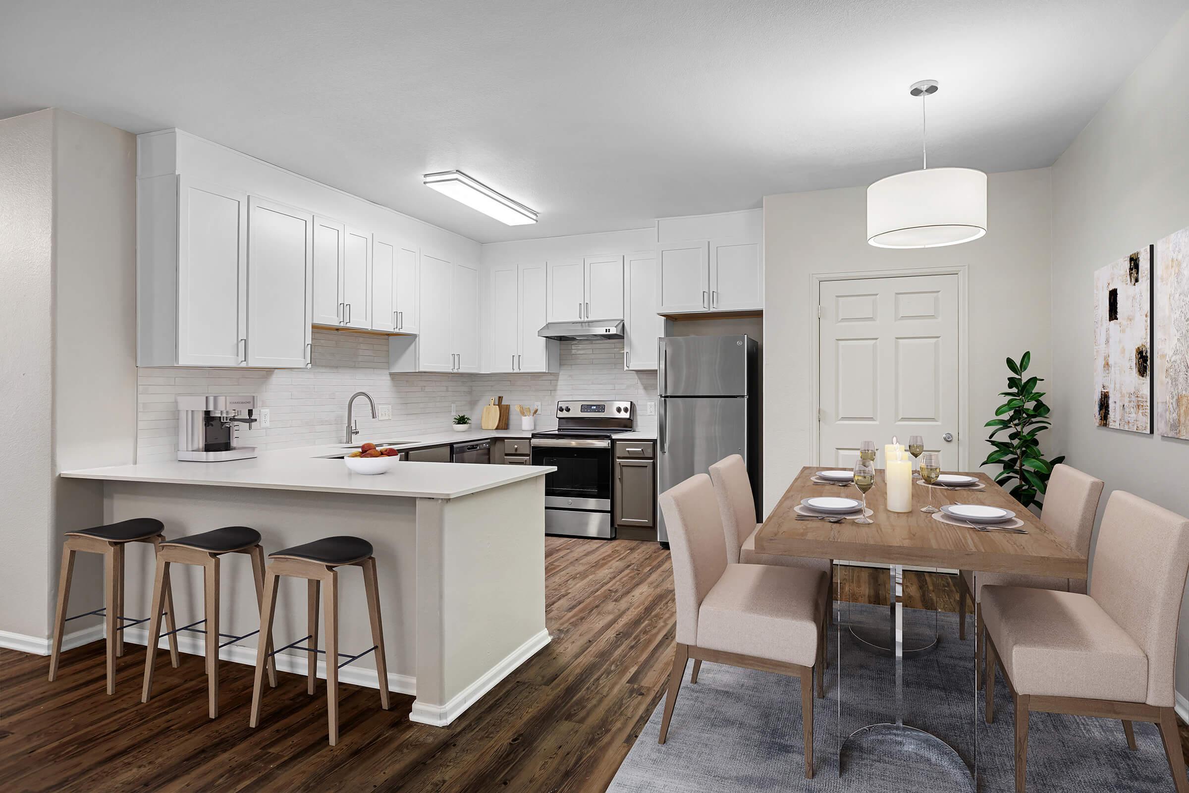 A modern kitchen and dining area featuring white cabinets, stainless steel appliances, a kitchen island with bar stools, and a wooden dining table with upholstered chairs. The space has warm wooden flooring and is brightly lit with overhead lighting and a pendant light above the dining table.