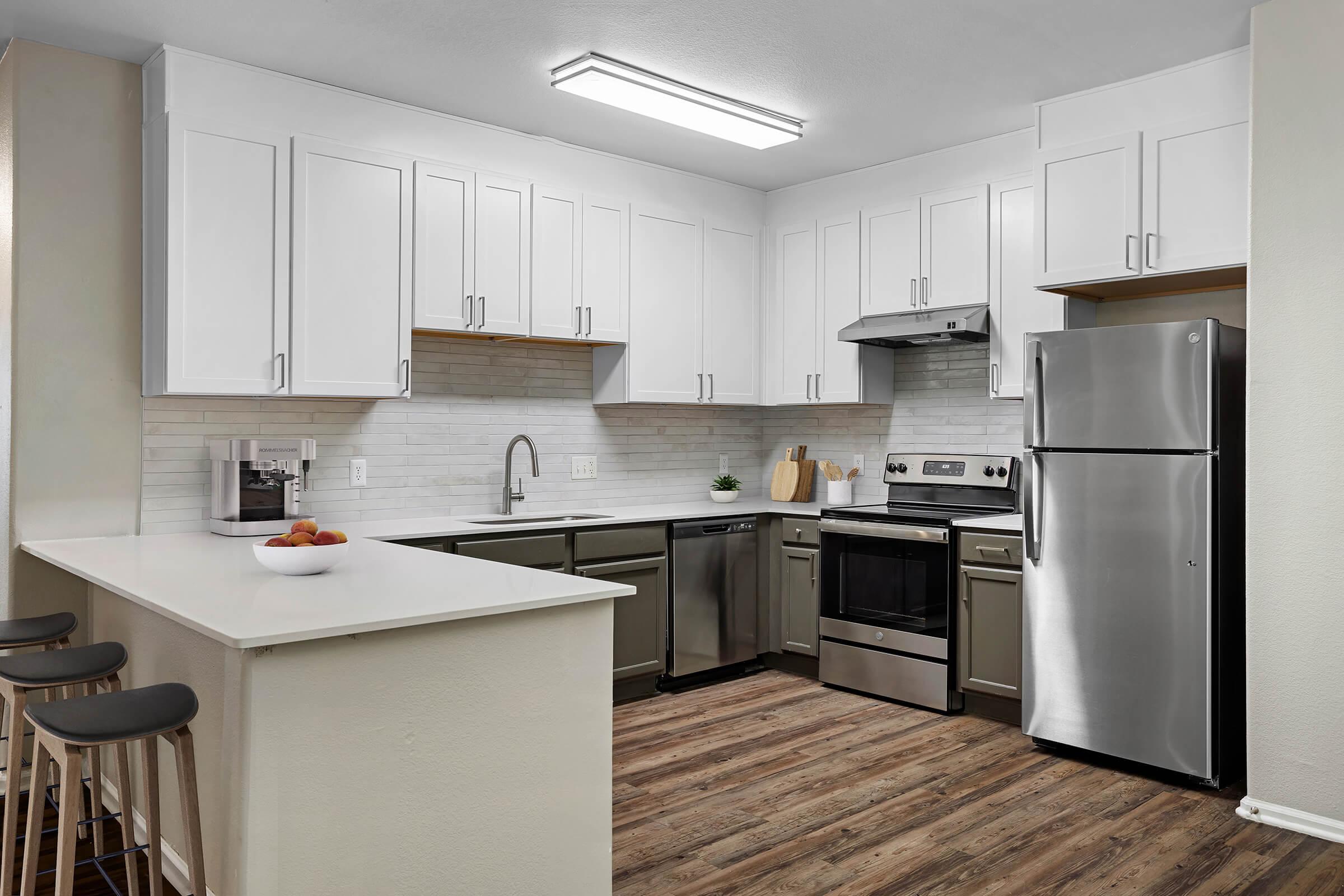Modern kitchen with white cabinetry and stainless steel appliances, including a stove, oven, and refrigerator. The countertop is light with a bar area featuring three wooden stools. A neutral-colored backsplash complements the cabinetry, and there's a small bowl of fruit on the counter. The flooring is wood-like laminate.