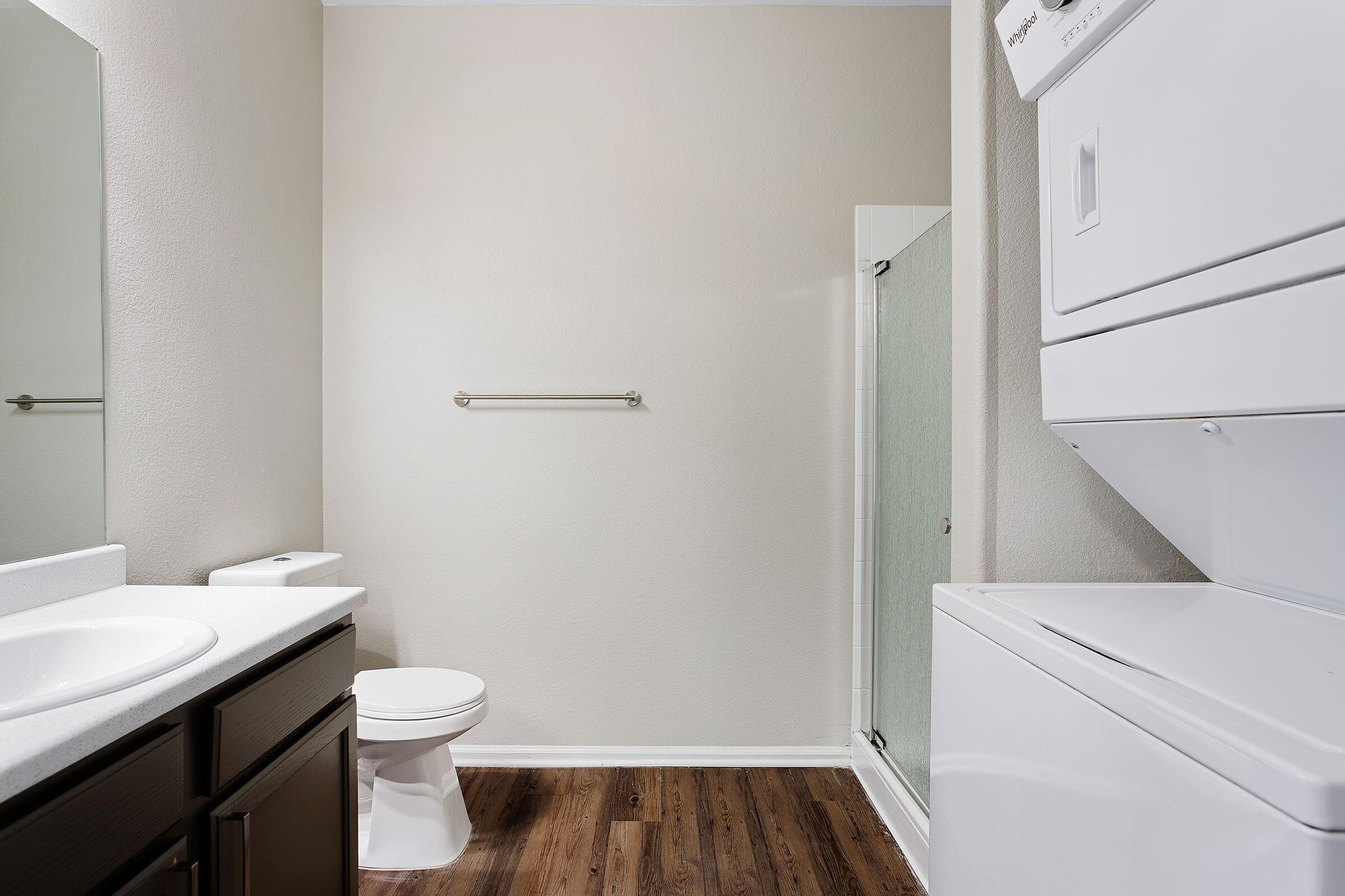 A clean, modern bathroom featuring a white sink and toilet, a glass shower stall, and a stacked washer and dryer. The walls are painted a light color, and the flooring is dark wood laminate. There is a towel bar on the wall opposite the shower.