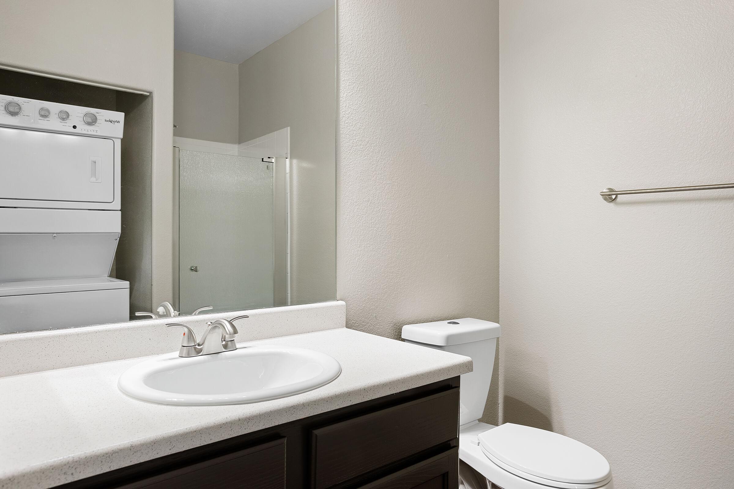A modern bathroom featuring a white sink with a chrome faucet, a toilet, and a washer/dryer unit stacked above each other. The walls are painted a neutral color, and there is a glass shower door visible in the background. A towel bar is mounted on the wall.