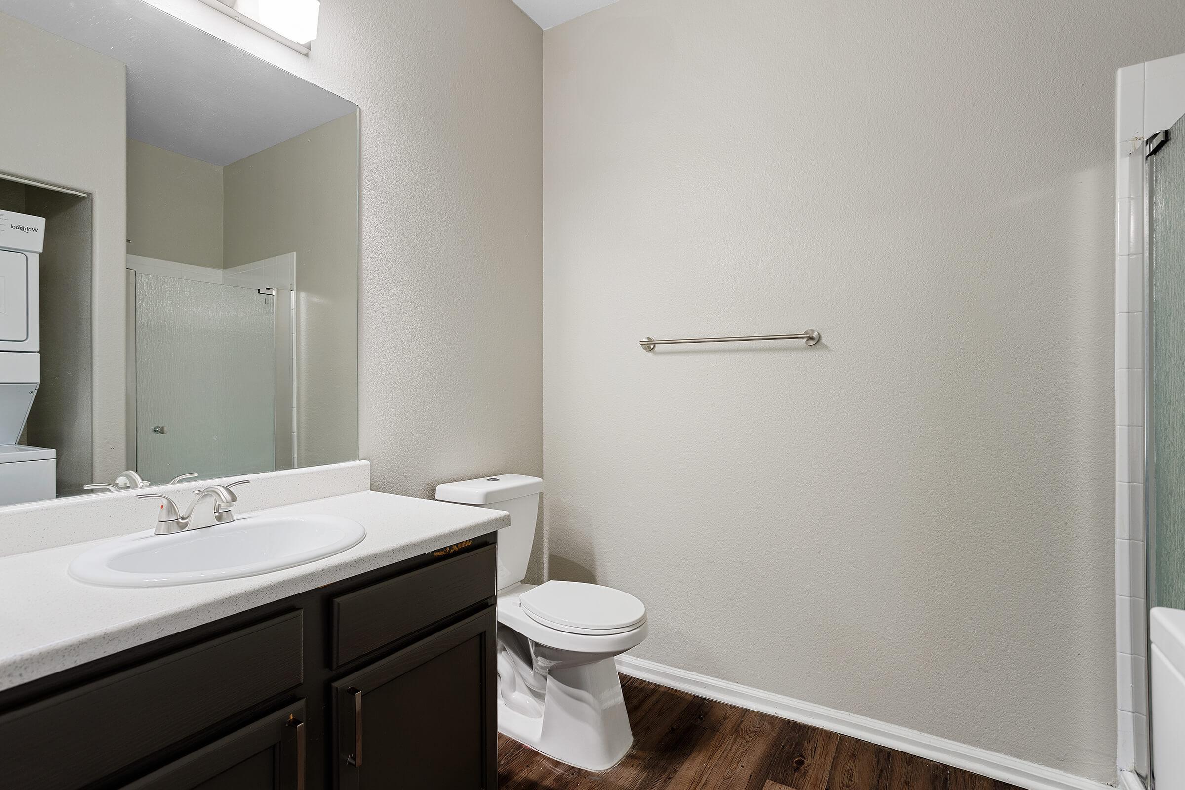 A clean, modern bathroom featuring a white sink with a mirror above, a toilet, and a towel rack. The walls are painted a light color, and there is a shower area visible. The flooring is wood-like, and a laundry area is partially visible in the background.