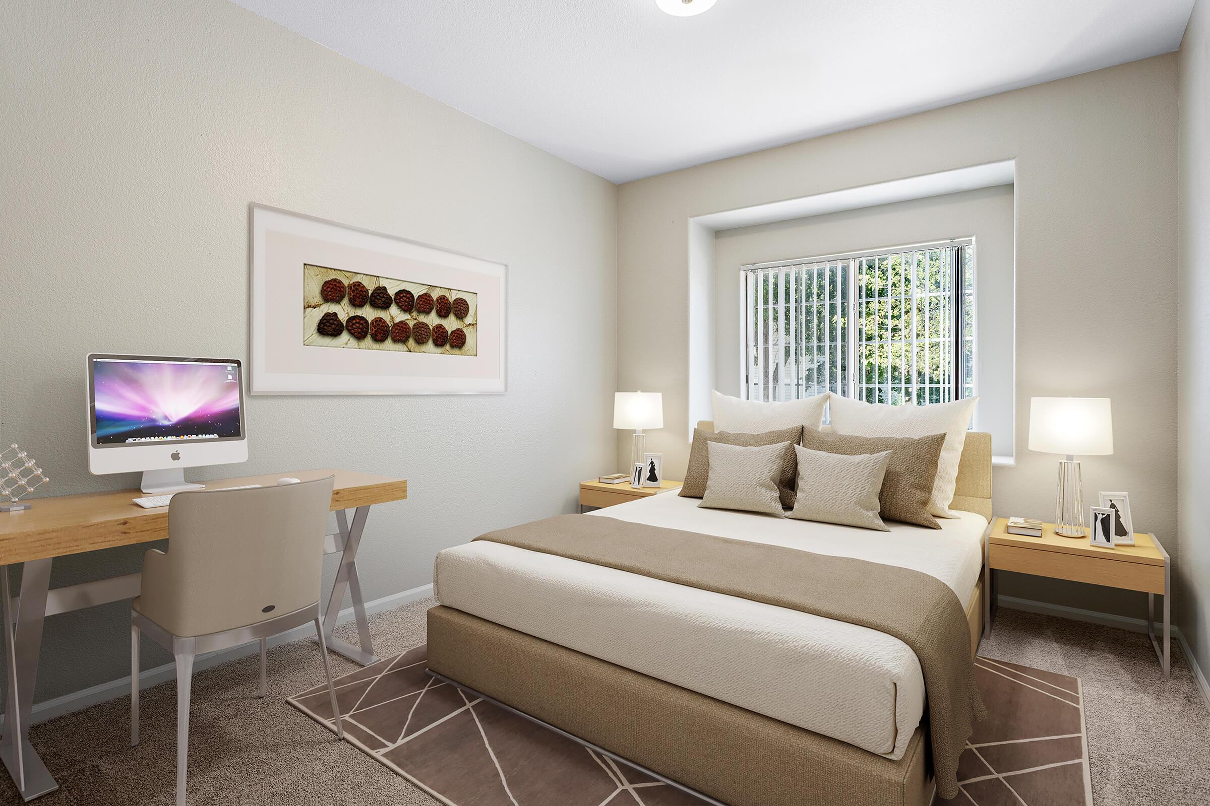 A cozy bedroom featuring a queen-sized bed with beige and cream bedding, two bedside lamps, a window with blinds, and an organized desk with a computer. The walls are painted light gray, and there is framed artwork above the bed, enhancing the warm and inviting atmosphere.