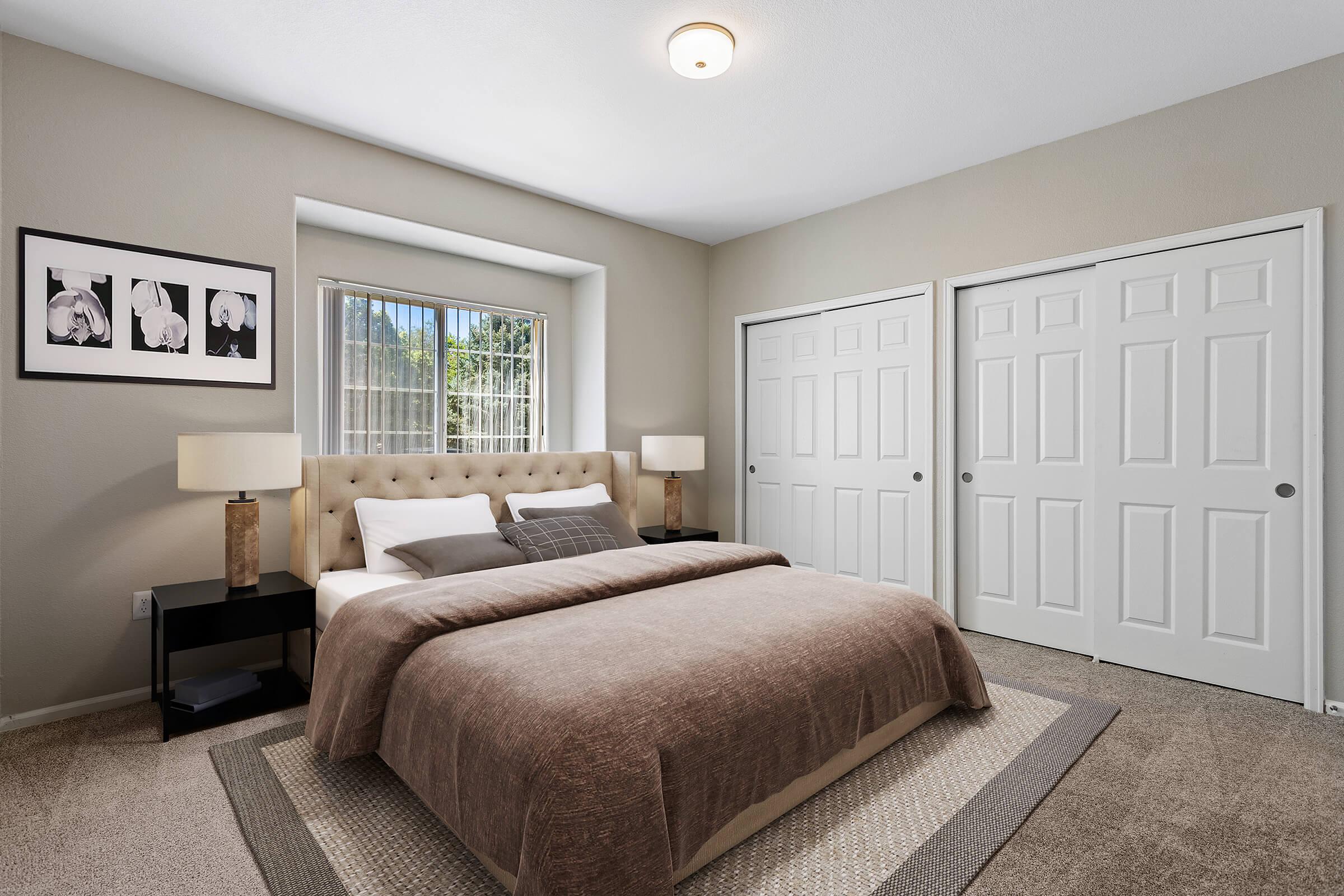A cozy bedroom featuring a queen-sized bed with a brown blanket, two bedside lamps, and a minimalist aesthetic. Wall art with floral prints is displayed above the bed. Natural light filters in through a window, and there are double closet doors visible on one side. The room has a neutral color palette and carpeted flooring.